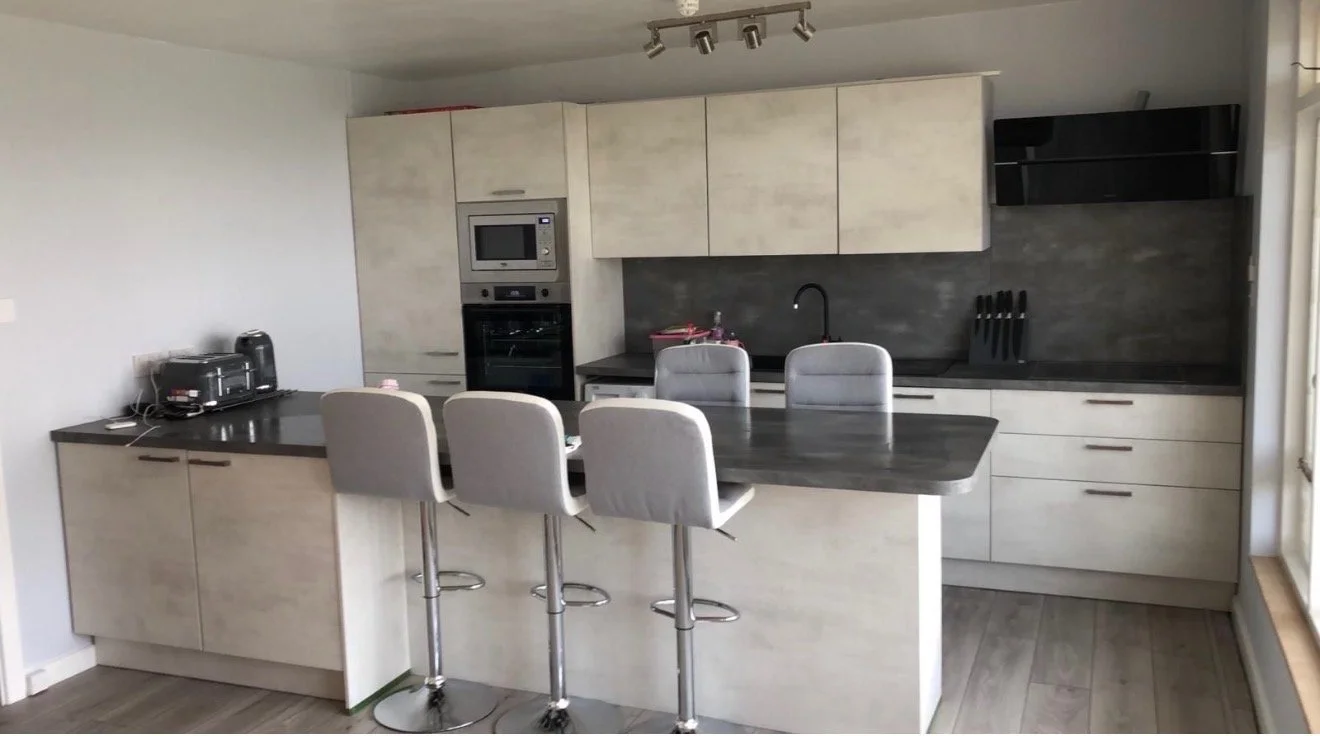 Modern kitchen with light-colored cabinets, gray countertops, and a dark gray backsplash. Four bar stools are at a kitchen island, which has a toaster and other small appliances. Built-in microwave and oven, sink with a faucet, and a set of knives ar
