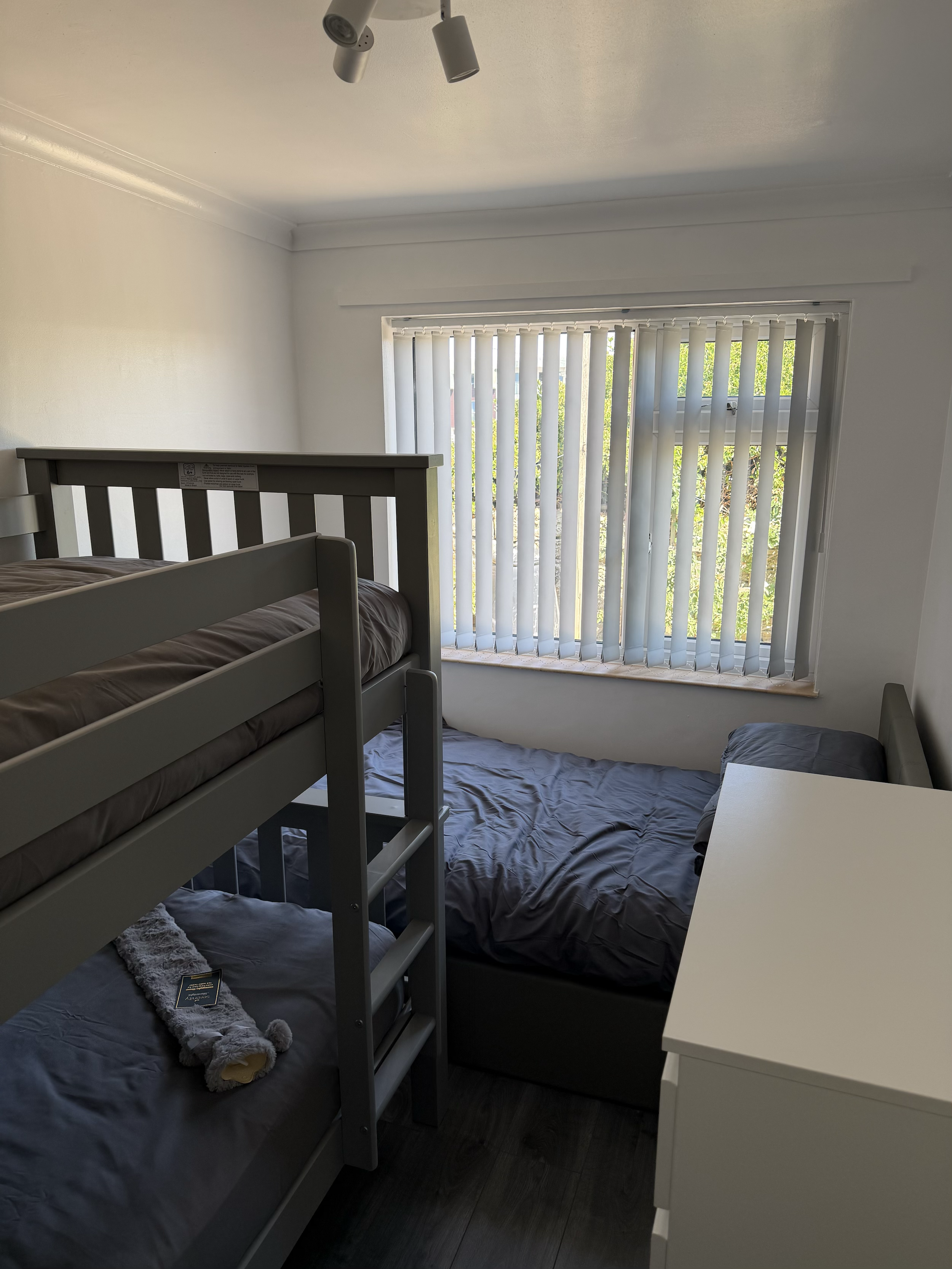 A small bedroom with white walls and a large window with vertical blinds, a gray bed with gray bedding, a white nightstand, and a gray bunk bed with a plush toy on top and a book or electronic device on the lower mattress.