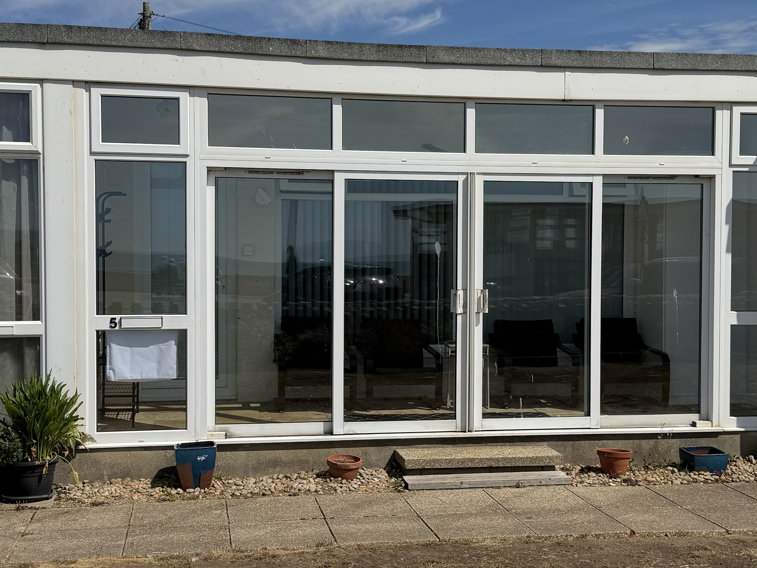 A white-framed glass conservatory with sliding doors, outside potted plants, and a gravel and stone pathway leading to the entrance.