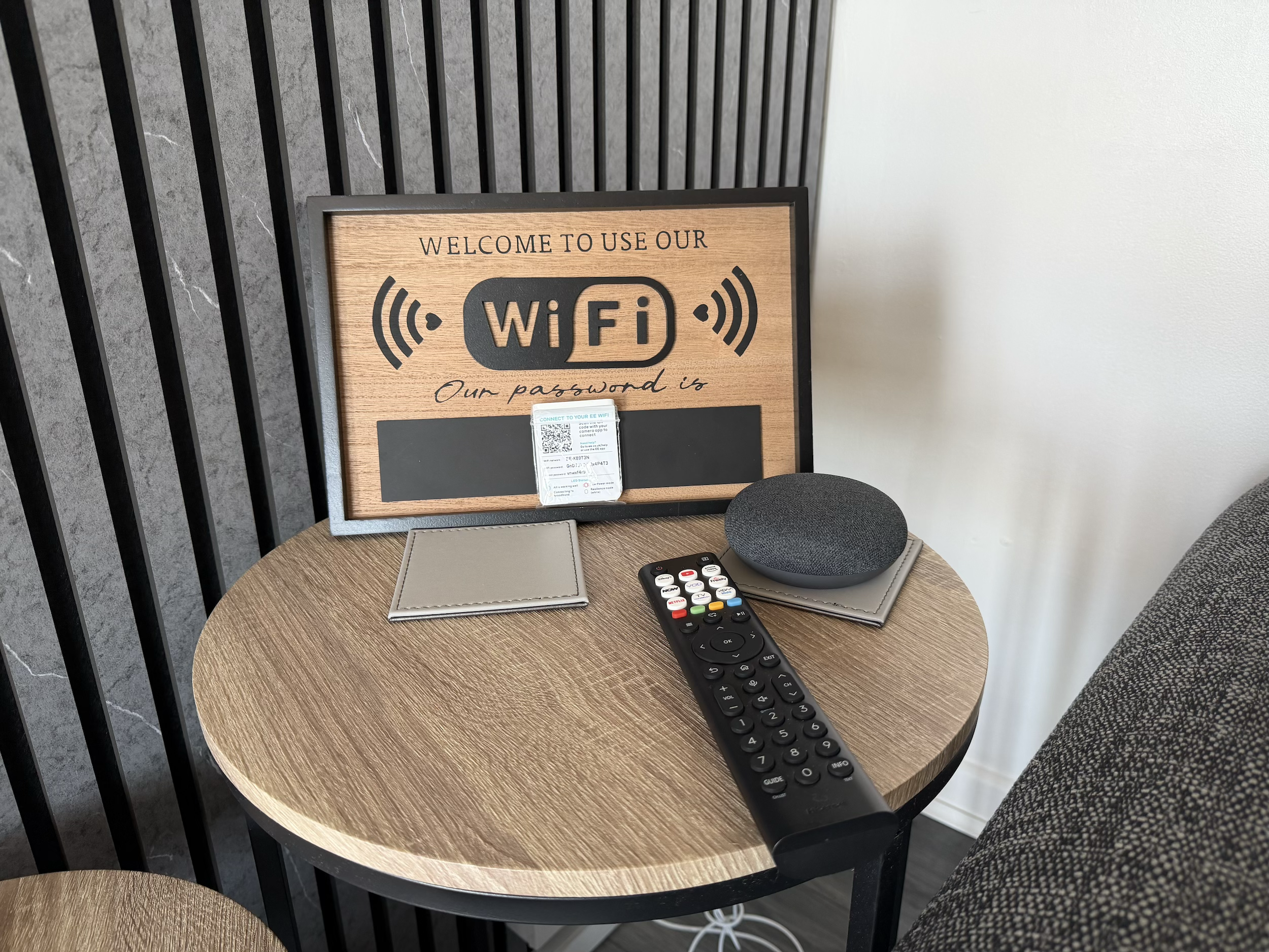 A round wooden table with a black TV remote, a gray notebook, a screen with Wi-Fi information and QR code, a gray speaker, and a beige object, next to a wall with a black slatted partition.
