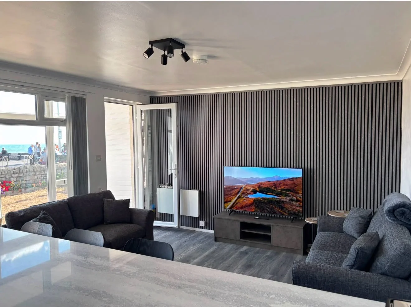 Modern living room with a gray and black color scheme, featuring a television on a gray entertainment center, gray sofas, a kitchen counter in the foreground, large sliding glass doors opening to an outdoor view of the beach and people, and a black ceiling light fixture.