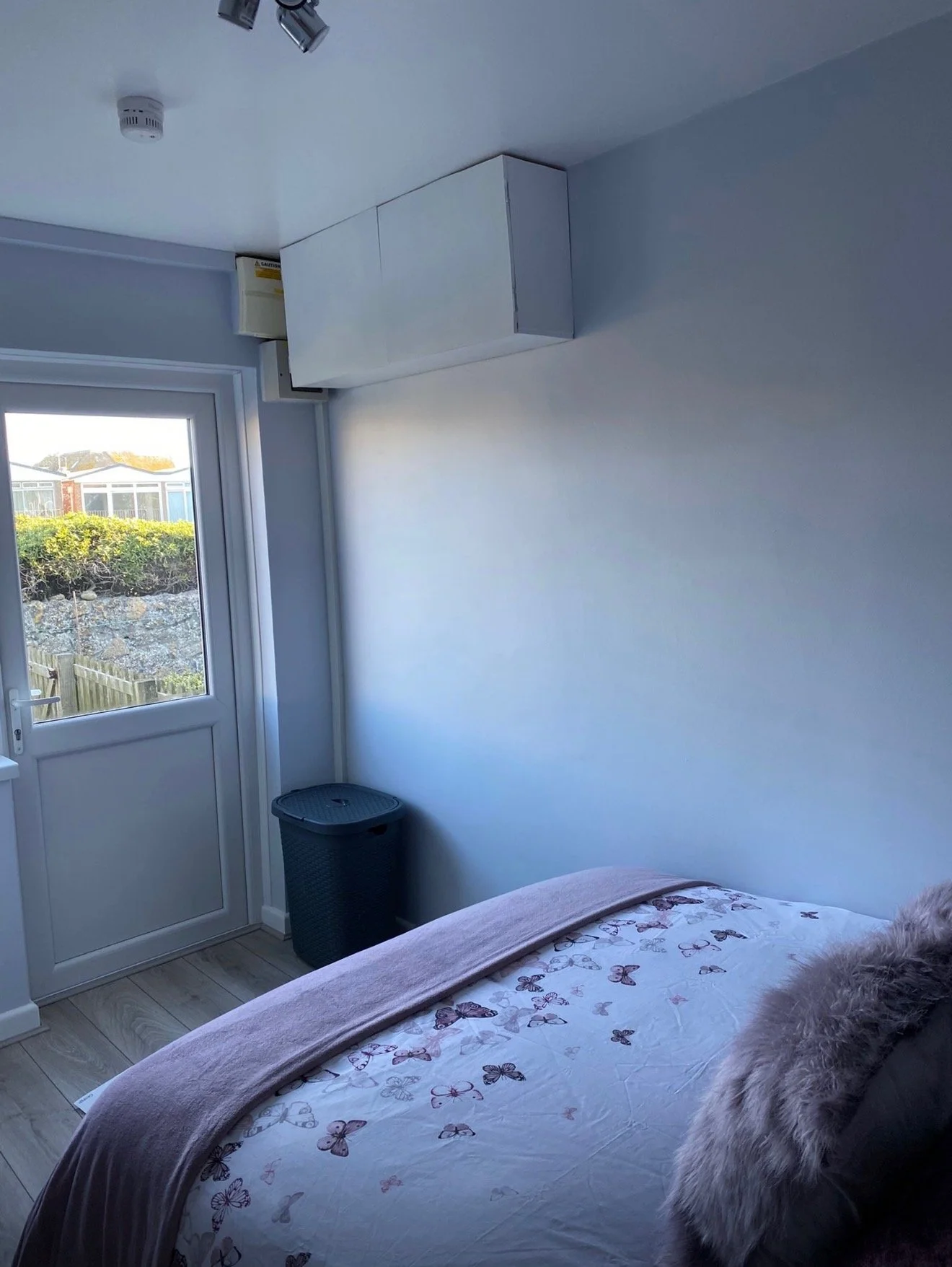 A bedroom with a bed covered in a butterfly-patterned duvet, a window with a view of a garden, a black laundry basket, and a white wall-mounted cabinet near the ceiling. Visible part of the room includes a door and ventilation and smoke detectors on 