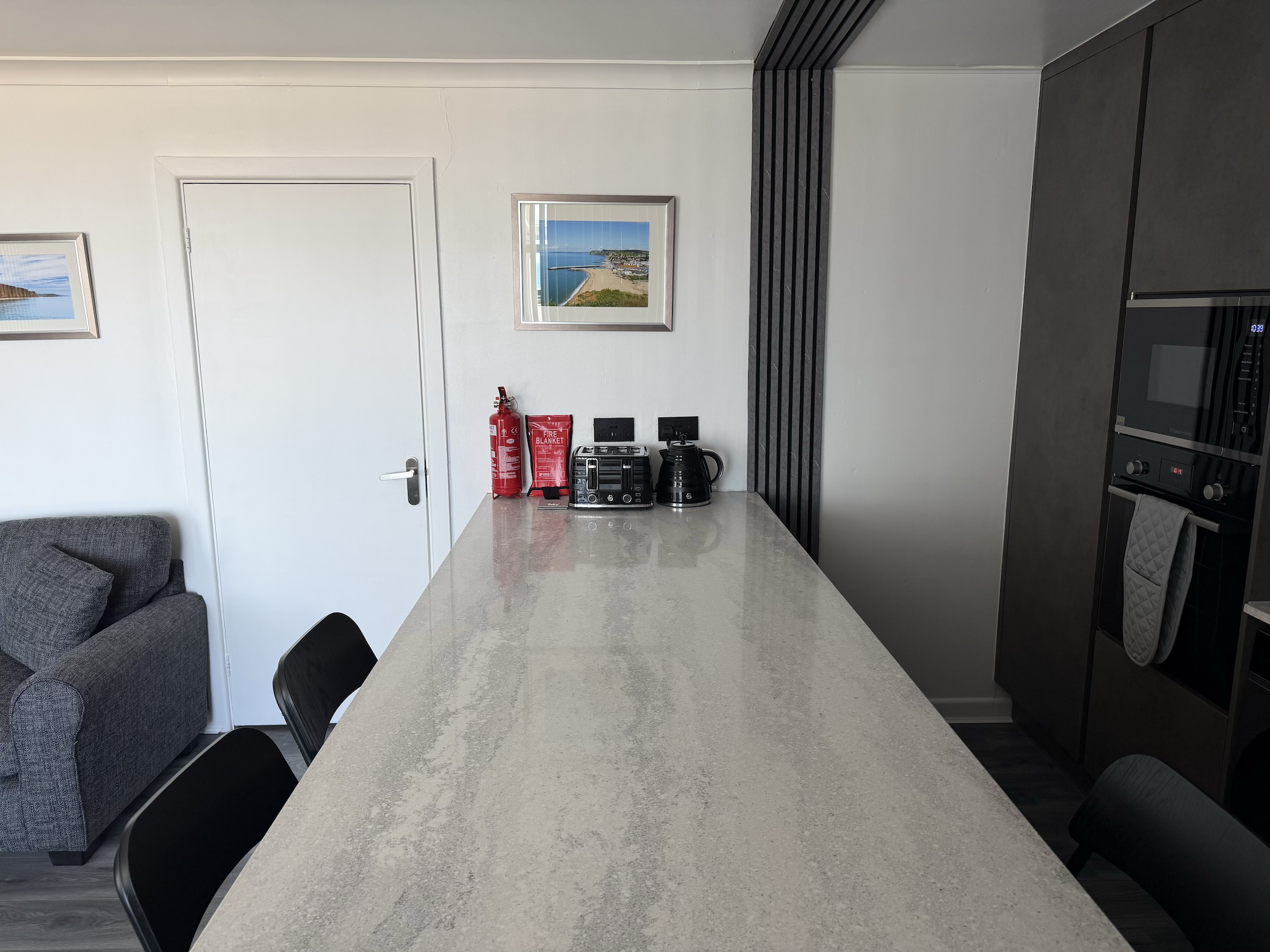 Modern kitchen with a white countertop, black chairs, and framed coastal landscape photographs on the white wall.