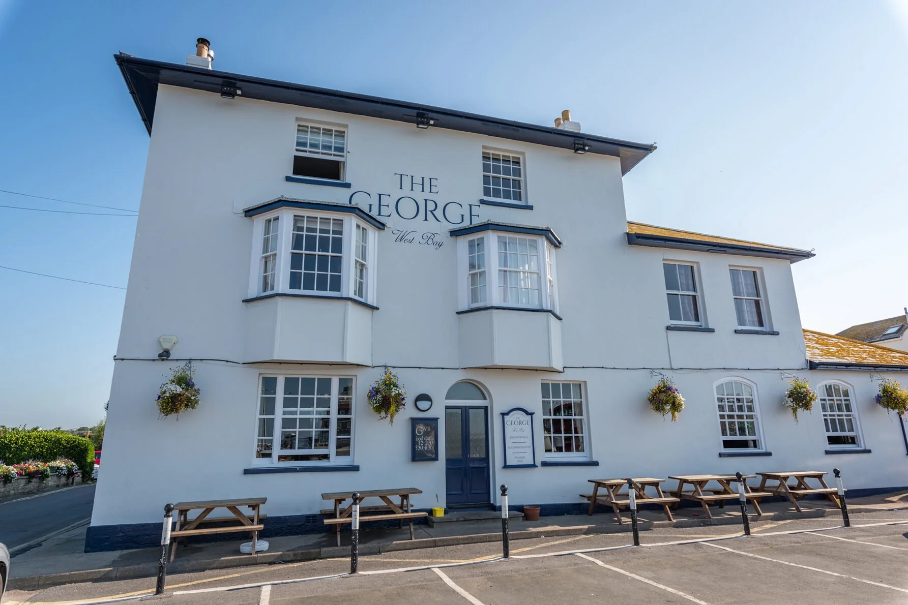 White building with blue window frames and trim, labeled 'The George West Bay'. Flower baskets hang outside, and there are outdoor benches in front. Clear sky above.