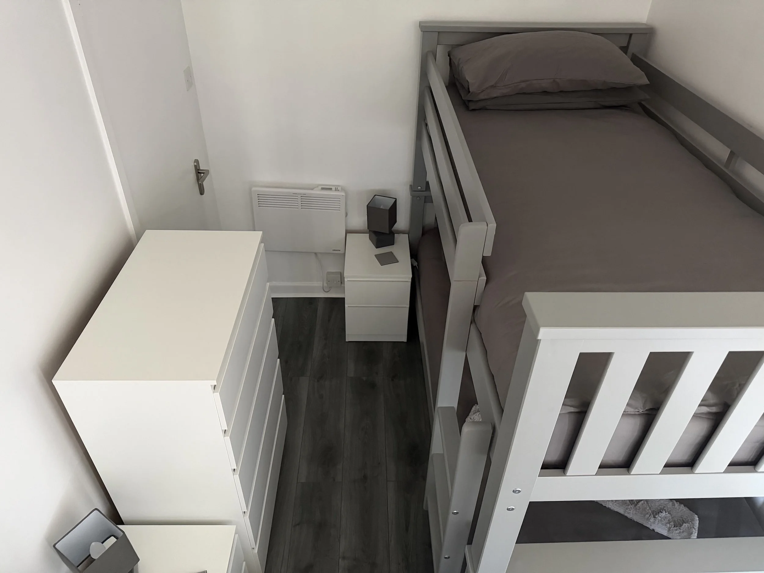 A bedroom with a white dresser, a gray bed frame, dark hardwood floors, a bed with gray bedding and pillows, and a white wall with a heating vent and a black speaker on the nightstand.