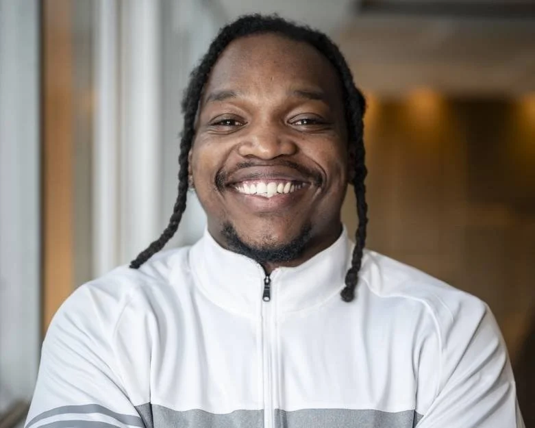 A smiling man with dreadlocks wearing a white sports jacket in an indoor setting.