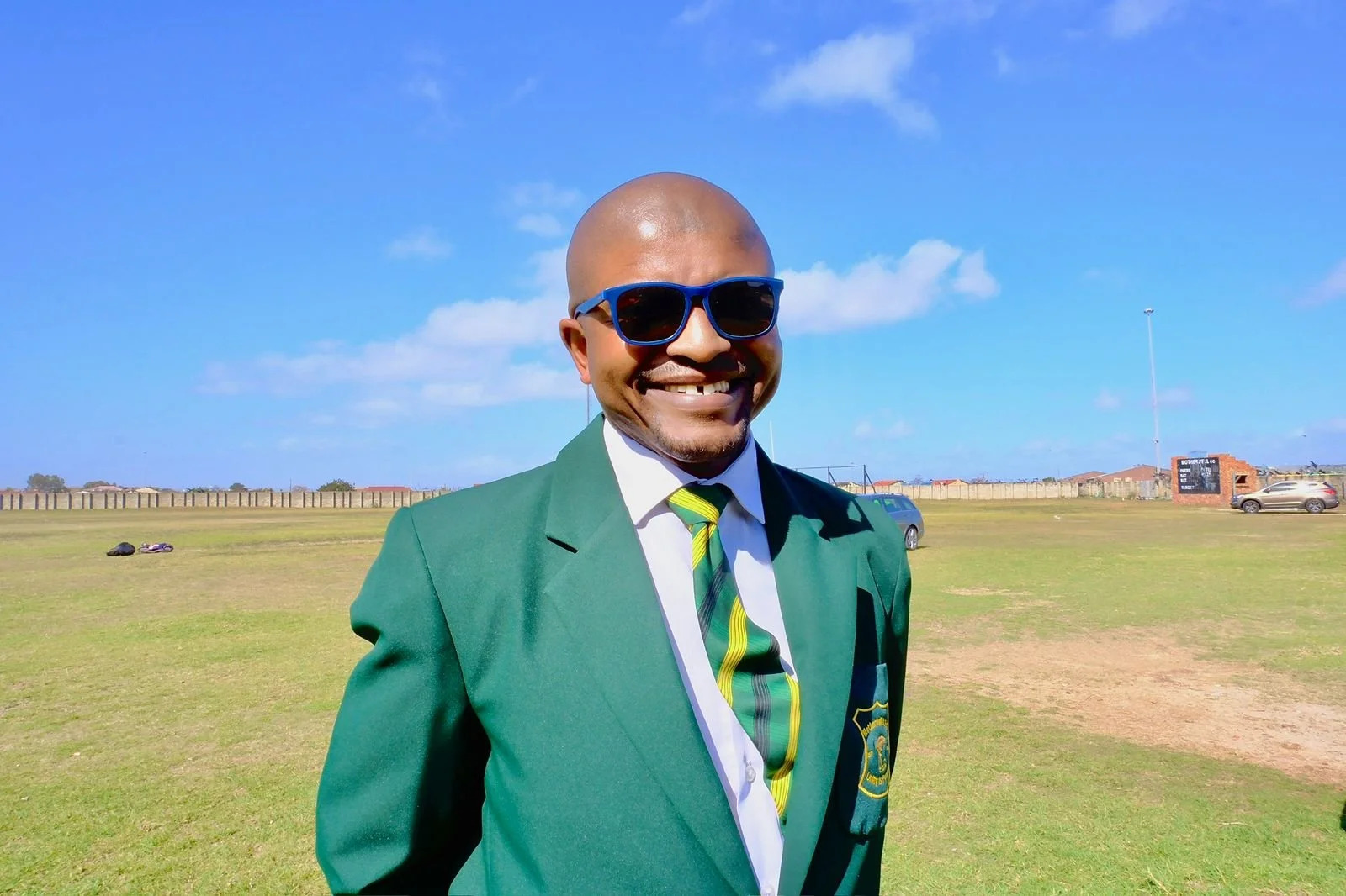 A man smiling outdoors wearing sunglasses, a green blazer, a white shirt, and a green and yellow striped tie, with a field, cars, and a blue sky in the background.