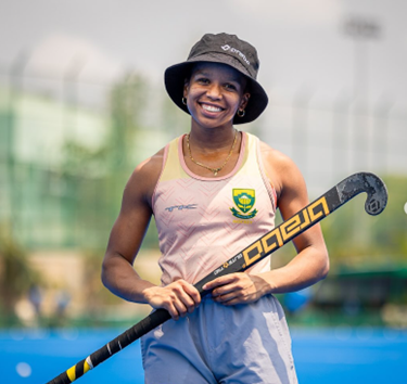 A young woman smiling and holding a field hockey stick on a field.