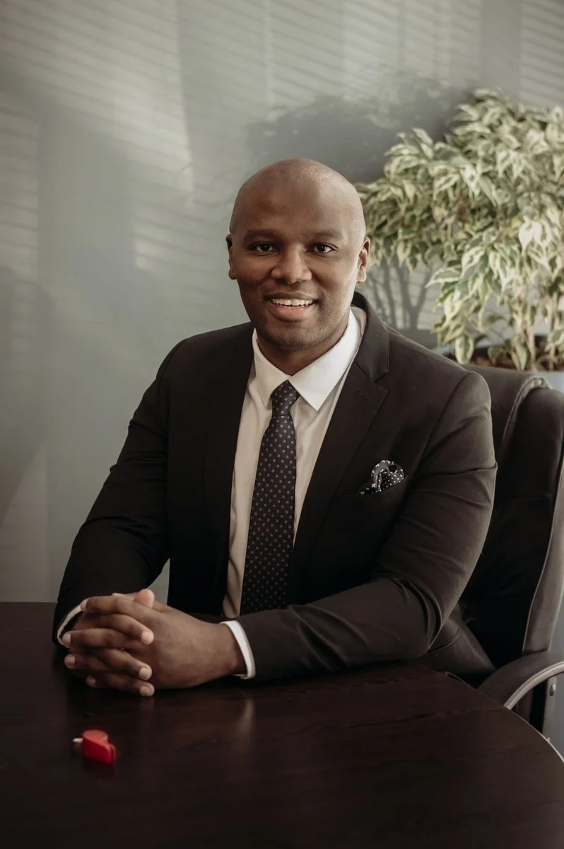 A professional man in a suit sitting at a dark wooden conference table, smiling, with a large houseplant and window blinds in the background.
