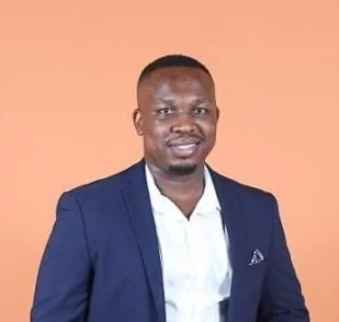 Portrait of a man in a dark blue suit and white shirt, standing against a peachy background.