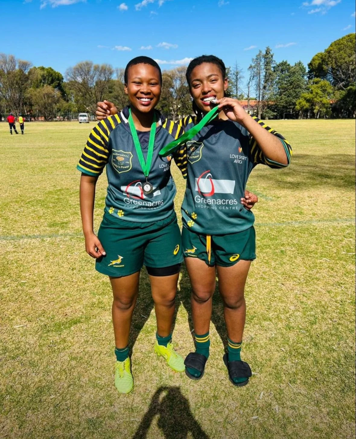 Two young women in green sports uniforms celebrating on a sunny field, one biting a medal, with trees and a blue sky in the background.