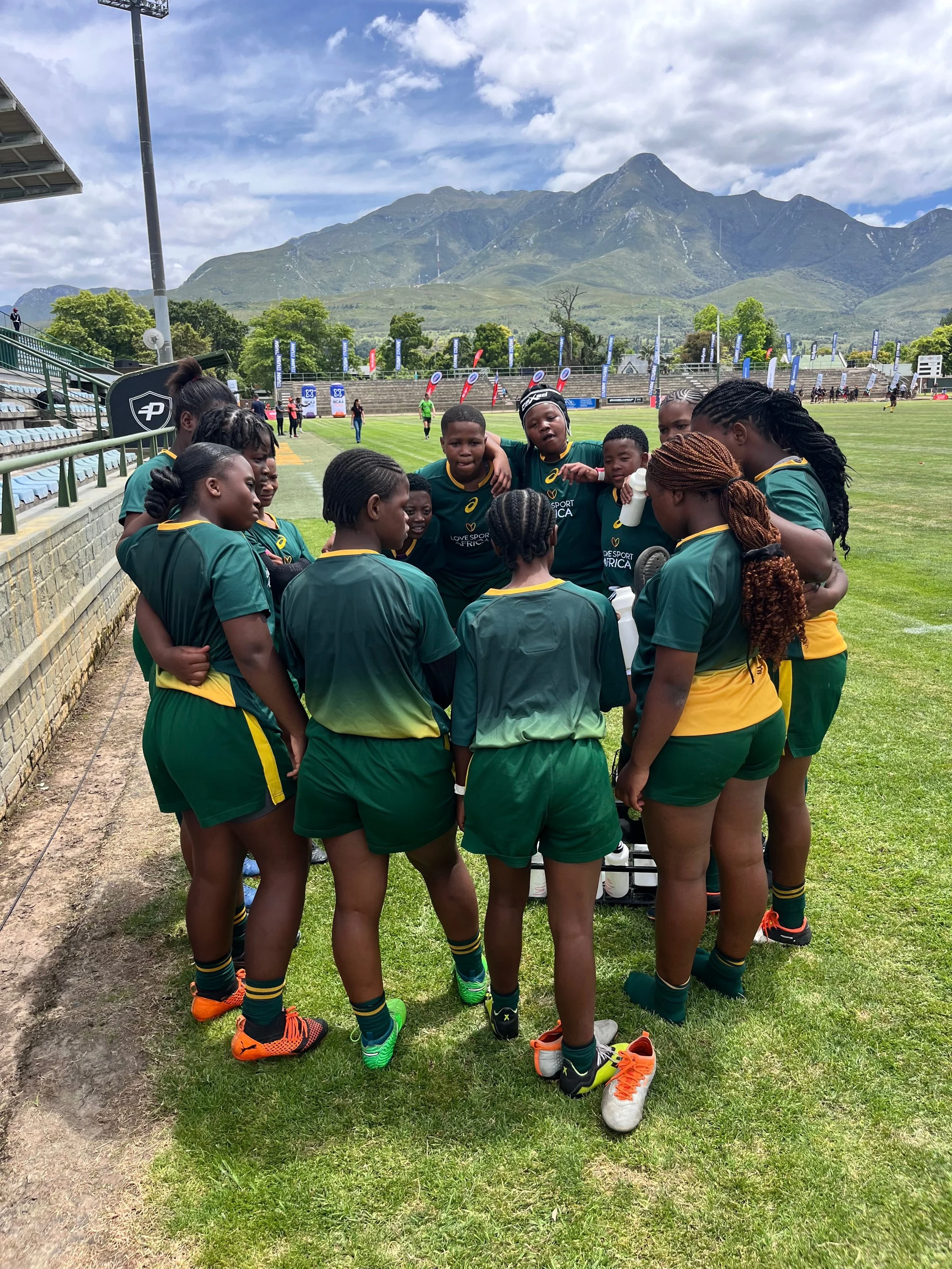 A group of young female rugby players huddled together on a rugby field, surrounded by mountains and a cloudy sky.