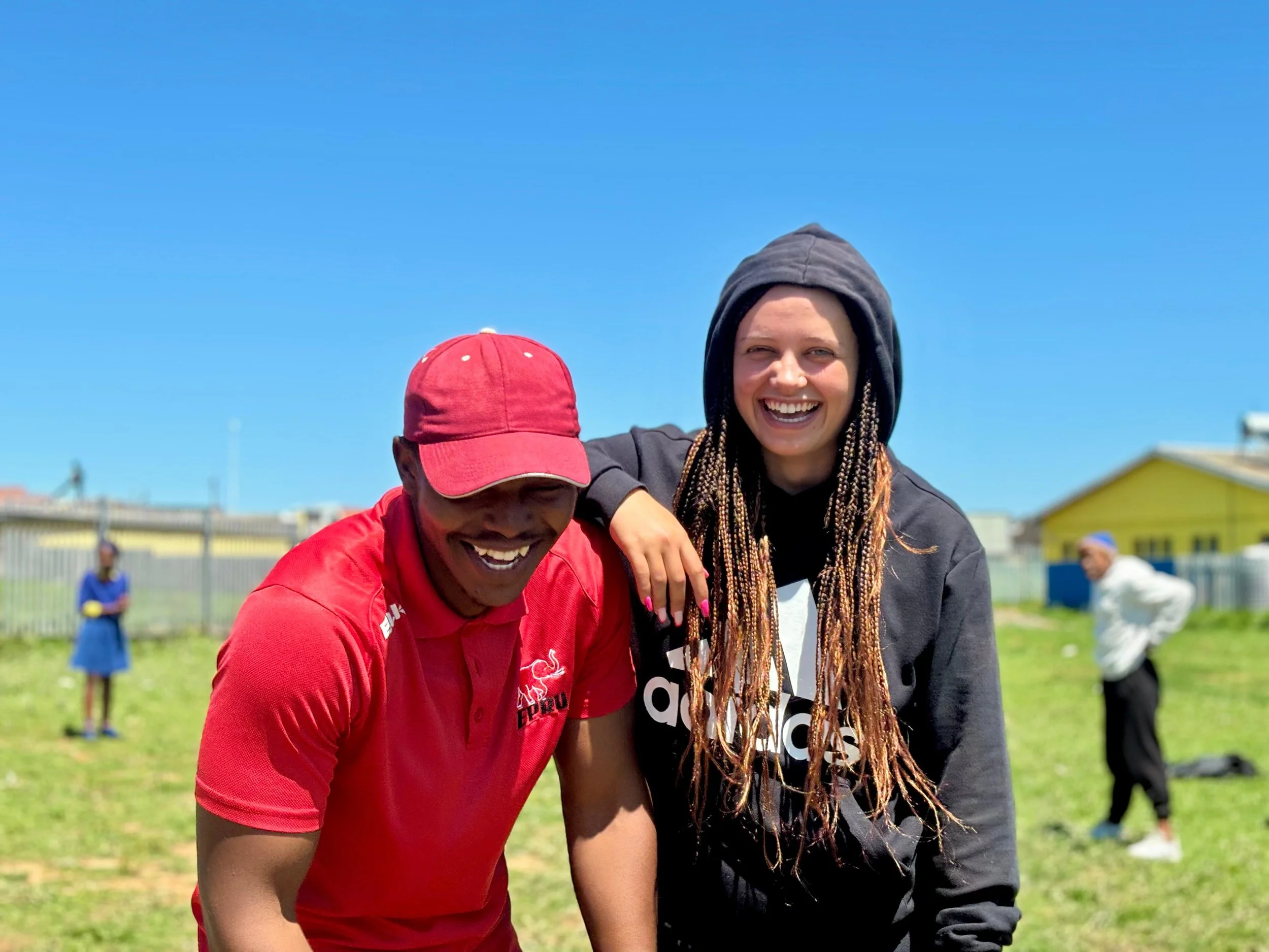 Two smiling people, one in a red cap and shirt and the other in a black hoodie, standing on grass under a clear blue sky with others in the background.