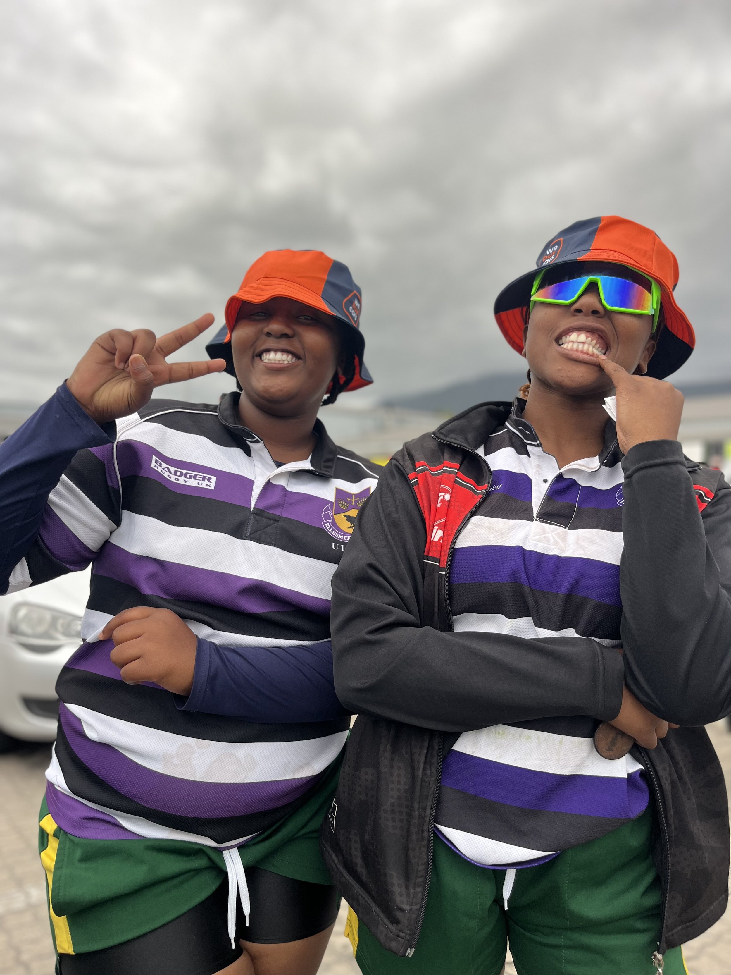 Two young women in sports uniforms and orange hats smiling and posing outdoors on a cloudy day.