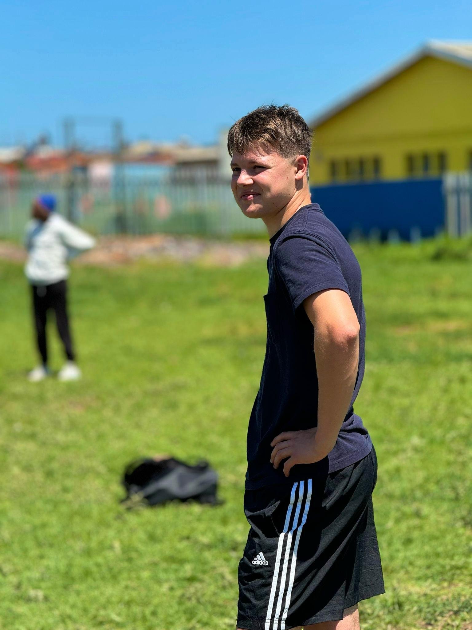 A young man standing with hands on hips on a grassy field, with a woman blurred in the background and a colorful building in the background.