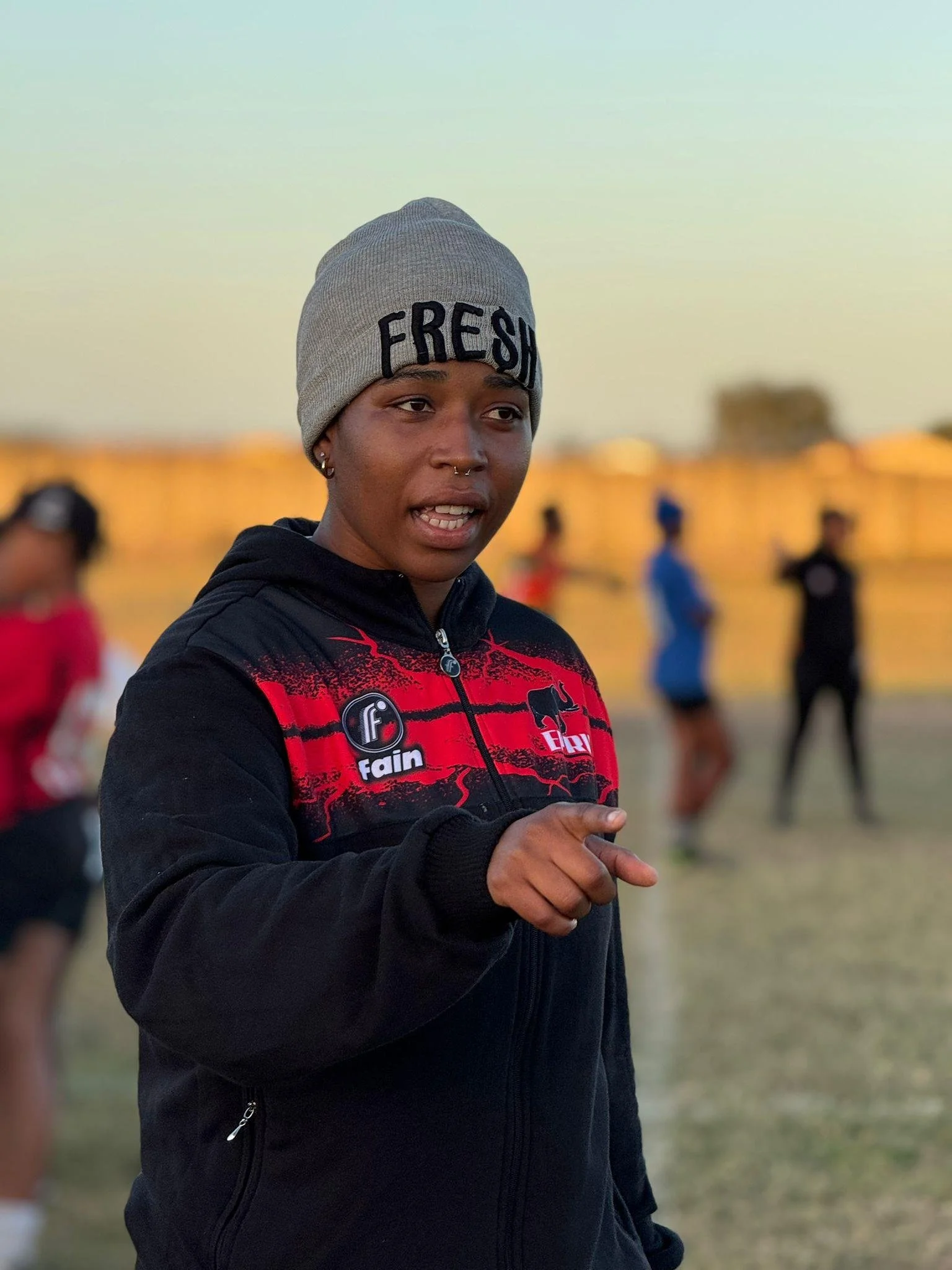 A young woman wearing a gray beanie with the word 'FRESH' on it and a black and red sports jacket, gesturing with her right hand on a grassy field during sunset, with other blurred people in the background.