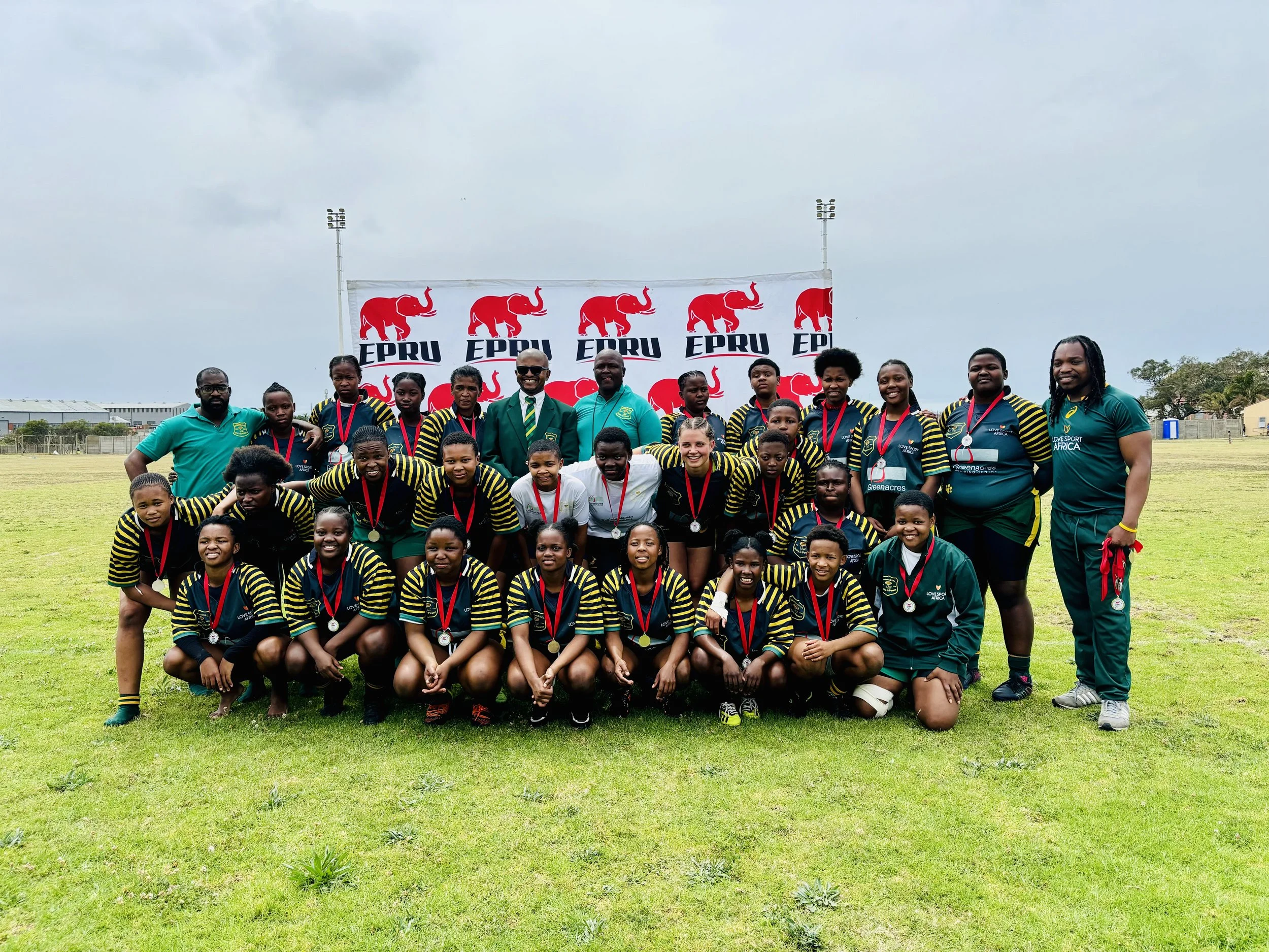 A group of young female athletes and their coaches posing for a team photo on a grassy field, wearing medals around their necks and sports uniforms with black and yellow stripes, in front of a banner with red elephants and the text 'EPRU'.