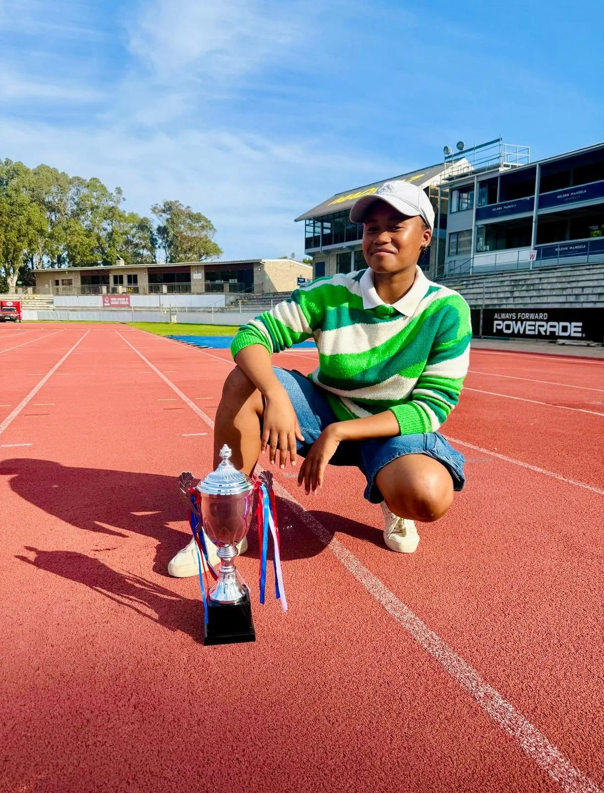 A young woman kneeling on a red running track with a trophy in front of her, wearing a white cap, green and white striped sweater, denim shorts, and white sneakers, smiling on a sunny day with a stadium and trees in the background.