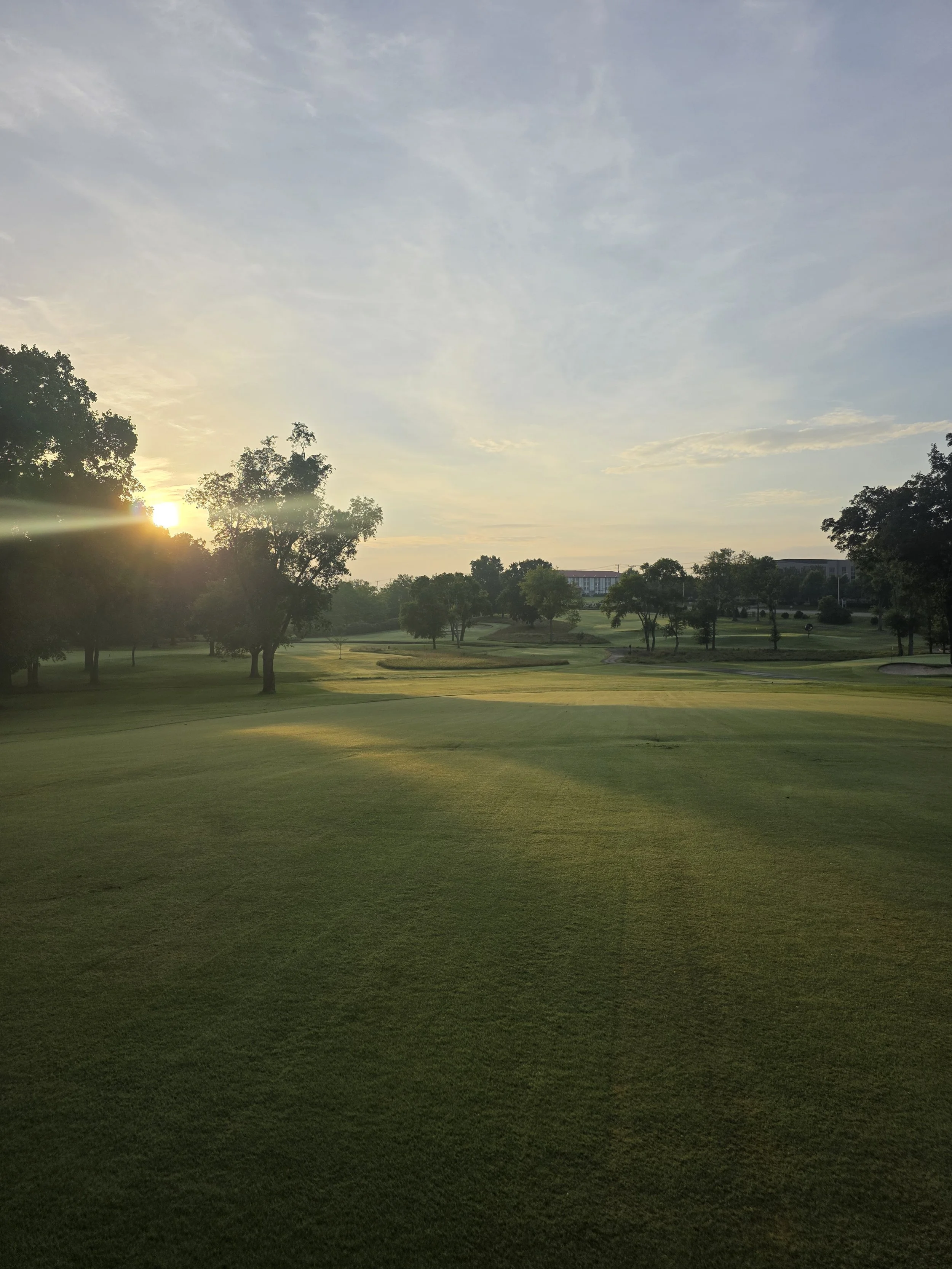 A golf course with lush green grass and several trees, with the sun setting or rising on the horizon, casting a warm glow across the sky and landscape.