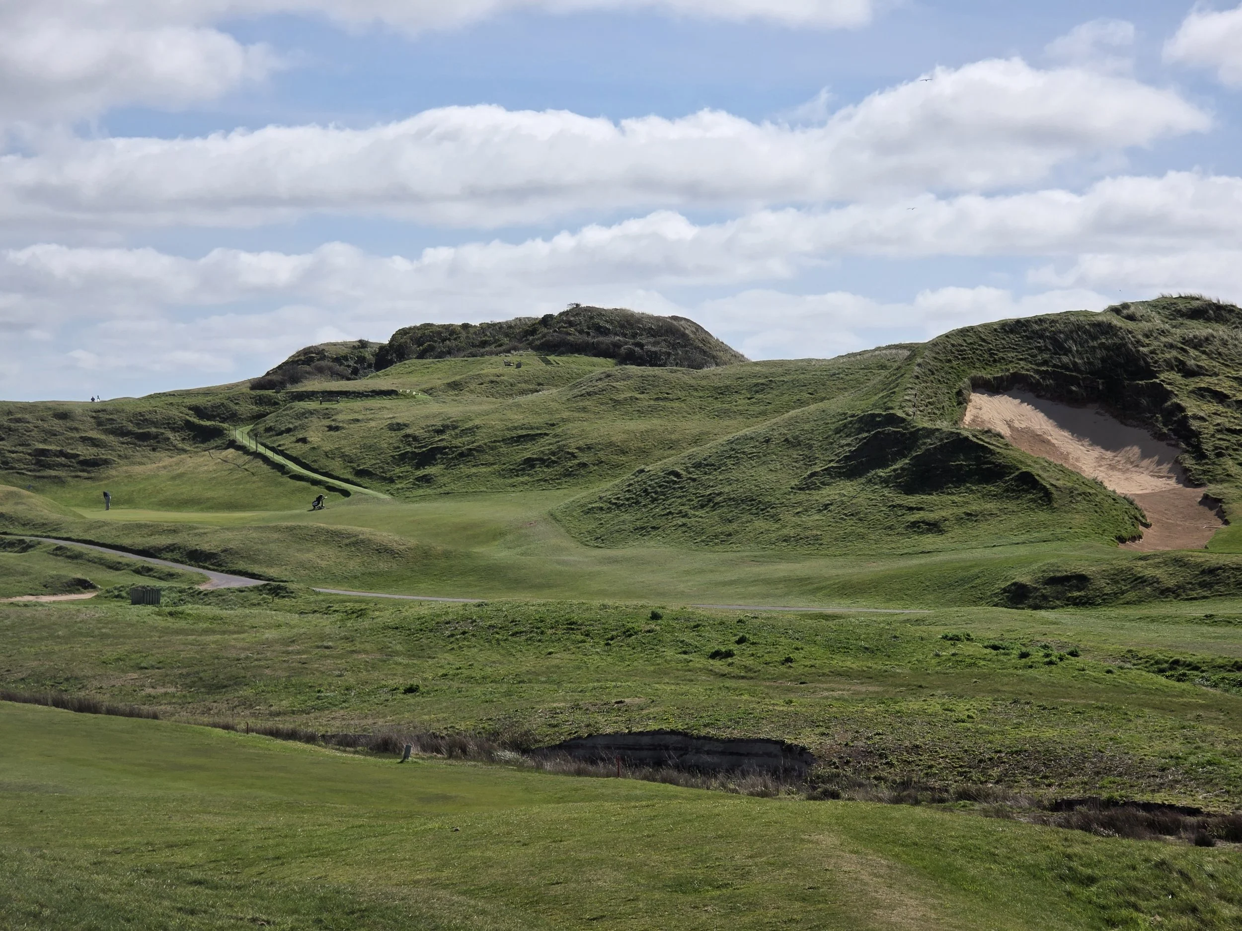 A scenic view of a golf course set in green, hilly terrain with sand bunkers and a few golfers in the distance under a partly cloudy sky.