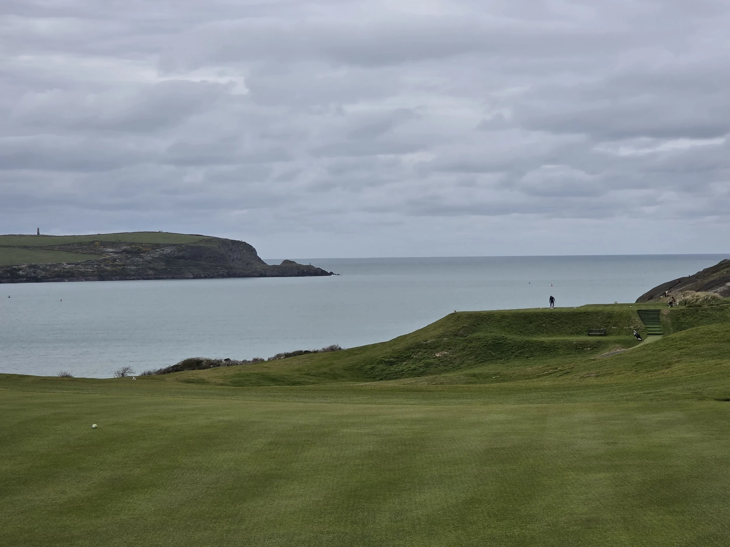 A golf course with a hole and a golf ball in the foreground, overlooking a body of water with cliffs and cloudy sky in the background.