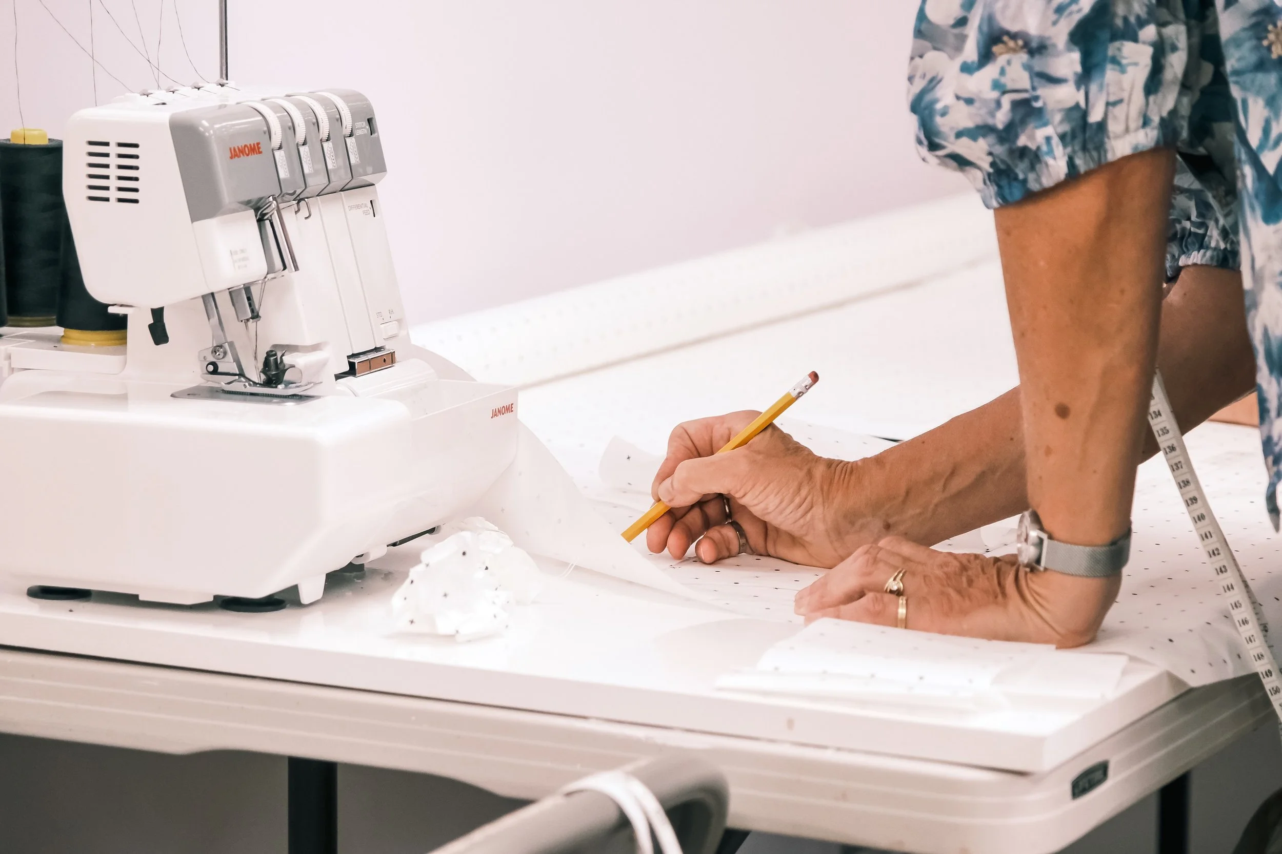 Close-up of a person measuring their arm with a measuring tape, with a sewing machine labeled 'Janome' in the background.