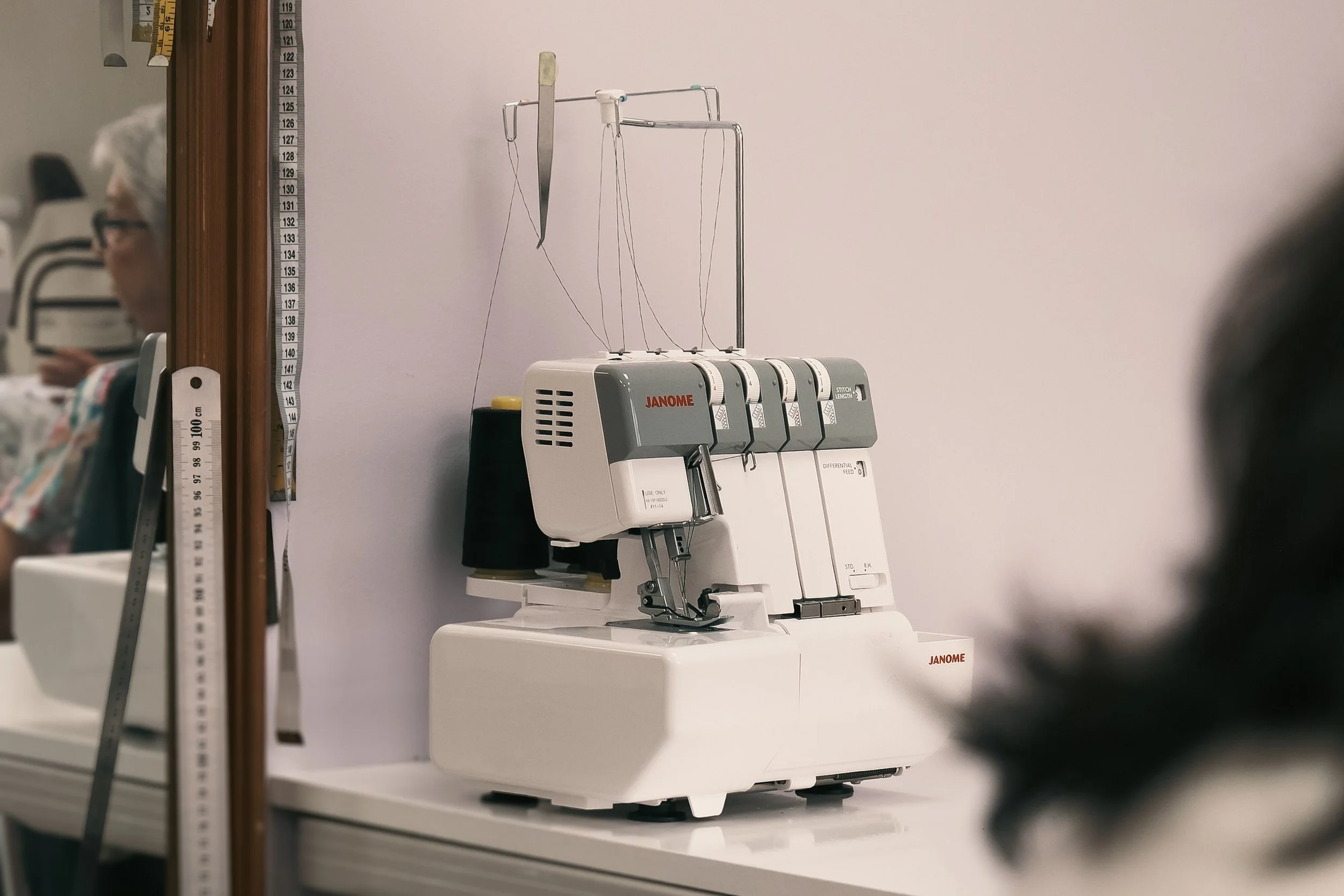 Close-up of a sewing machine, labeled 'JANOME,' with spools of thread and a metal stand in the background. A measuring tape is visible to the left, and a person with glasses and white hair appears in the blurred background.
