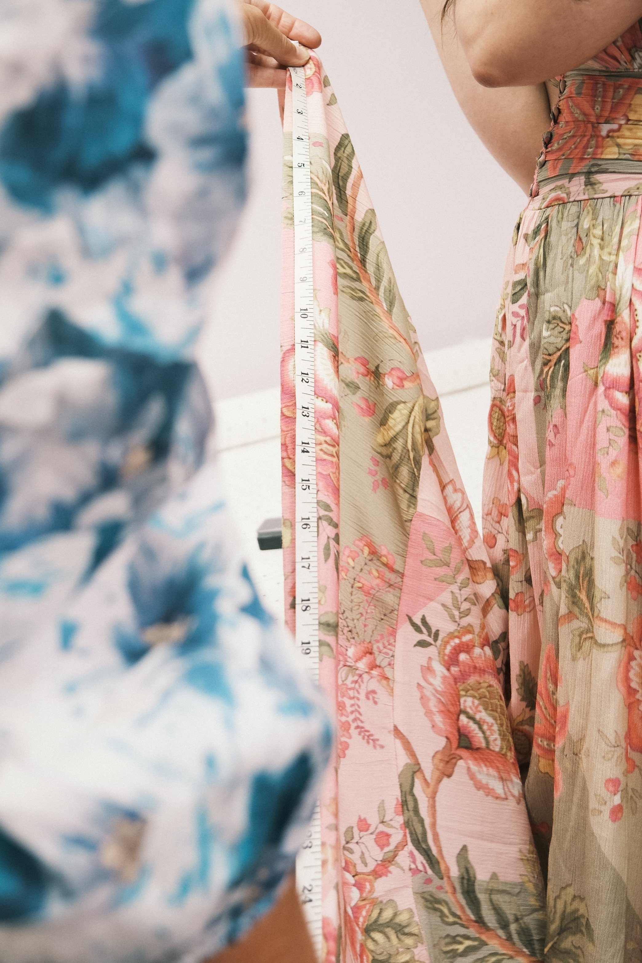 Close-up of a person measuring a pink floral dress with a measuring tape, while another person holds a piece of blue and white patterned fabric.