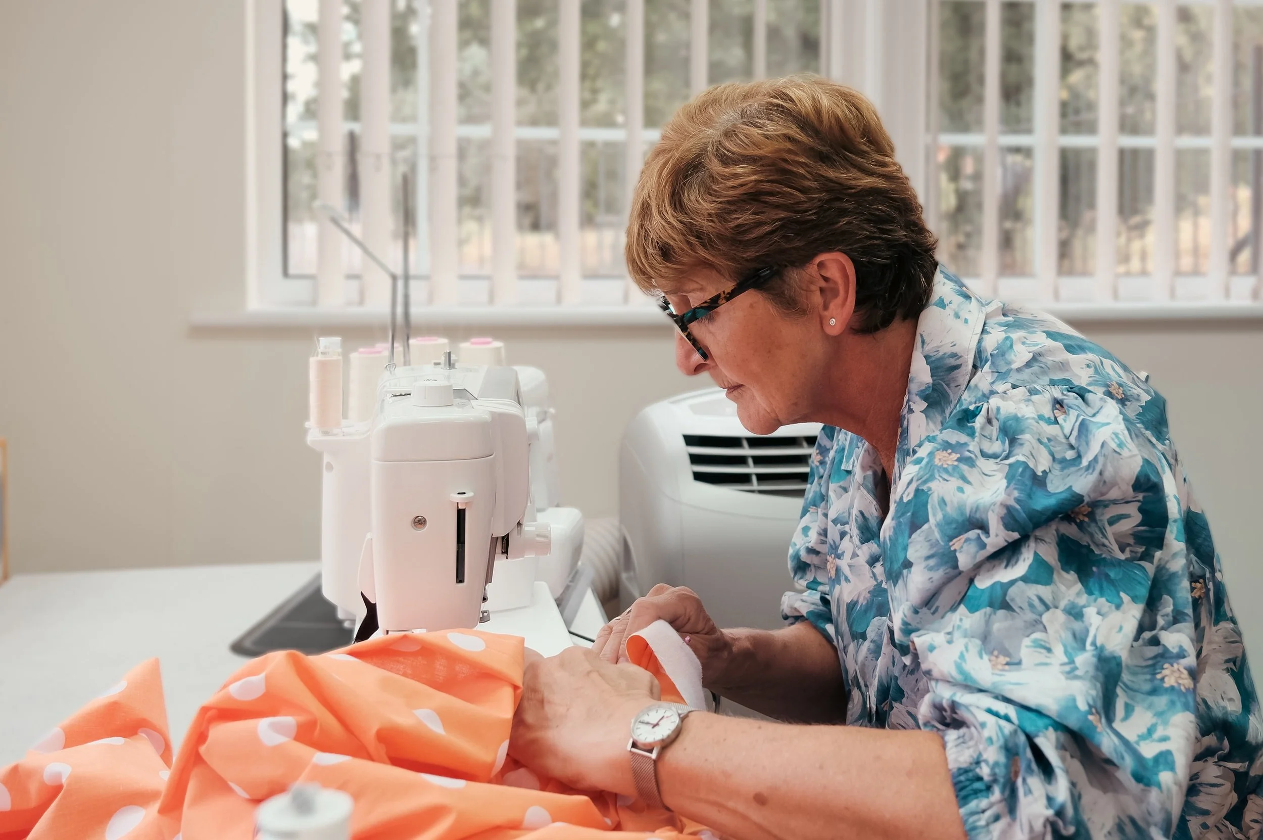 A woman with short, light brown hair and glasses working with a sewing machine on orange fabric with white polka dots in a well-lit room with large windows.