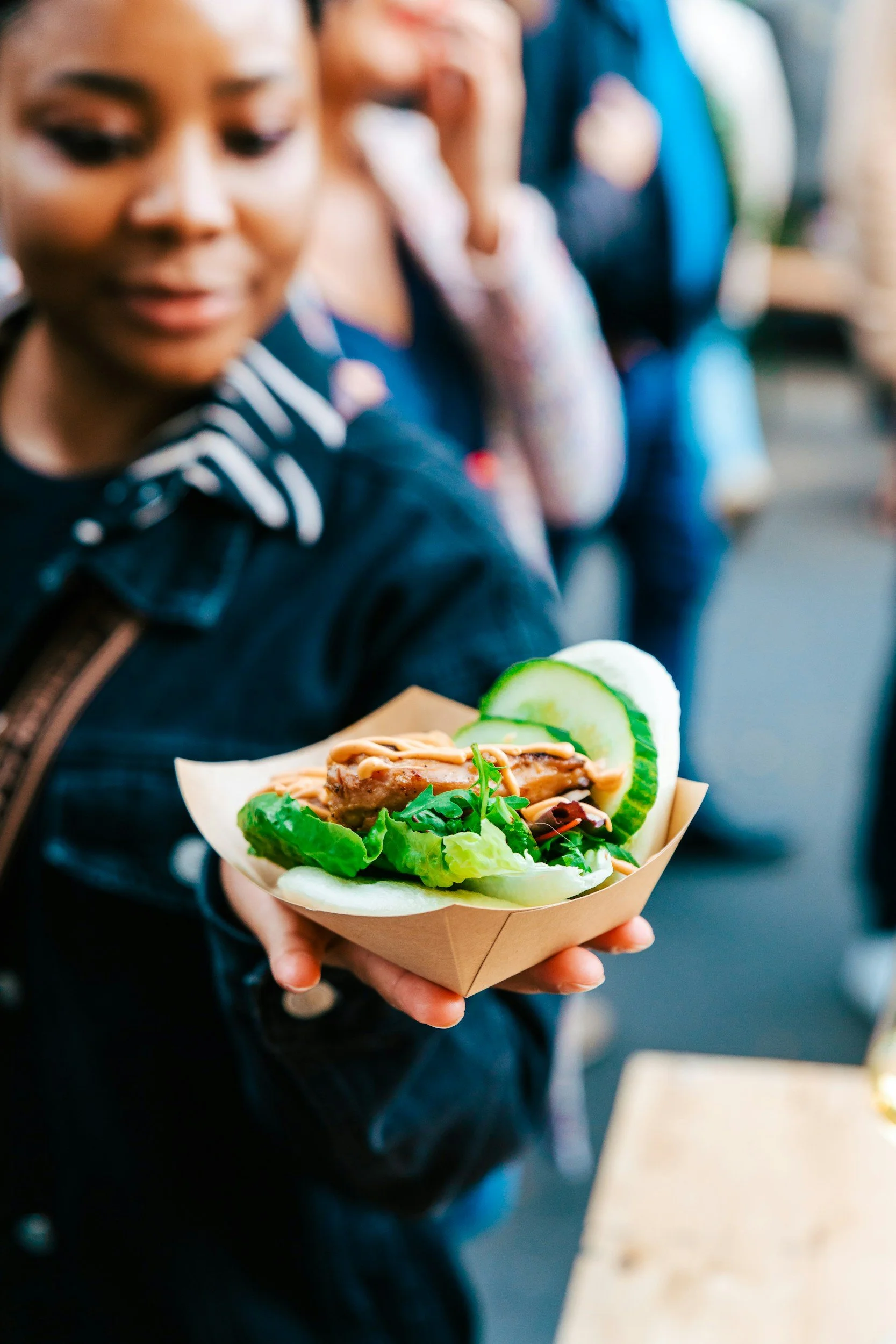 Close-up of a woman holding a food cone with sliced cucumber, leafy greens, and grilled meat. She is wearing a dark jacket with a striped collar. The background shows other people in a line or gathering.