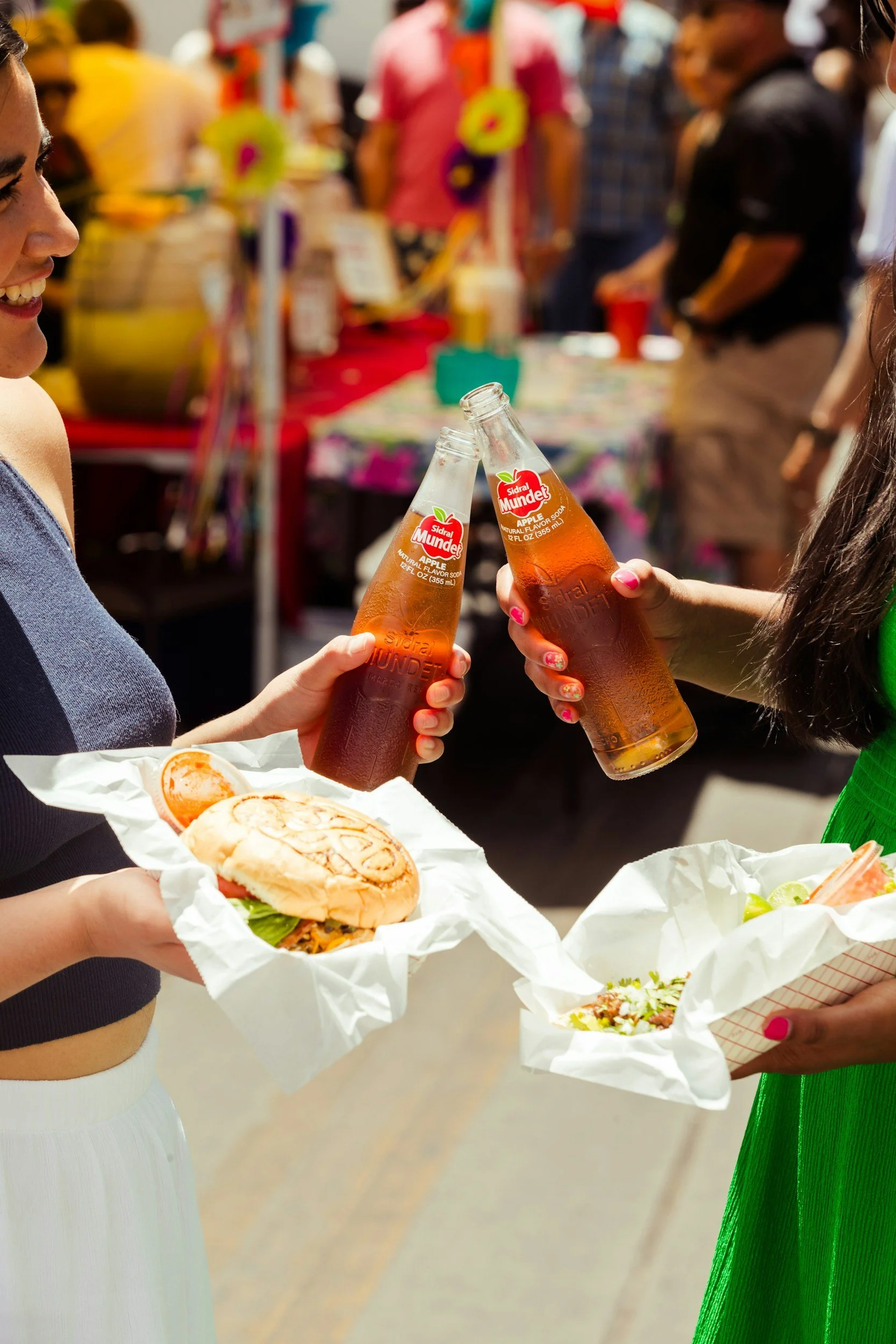 Two women are holding bottled sodas and trays of food at an outdoor event. One woman is holding a sandwich with lettuce, tomato, and meat, while the other holds a bowl of salad. In the background, there are several people and a table with colorful decorations.