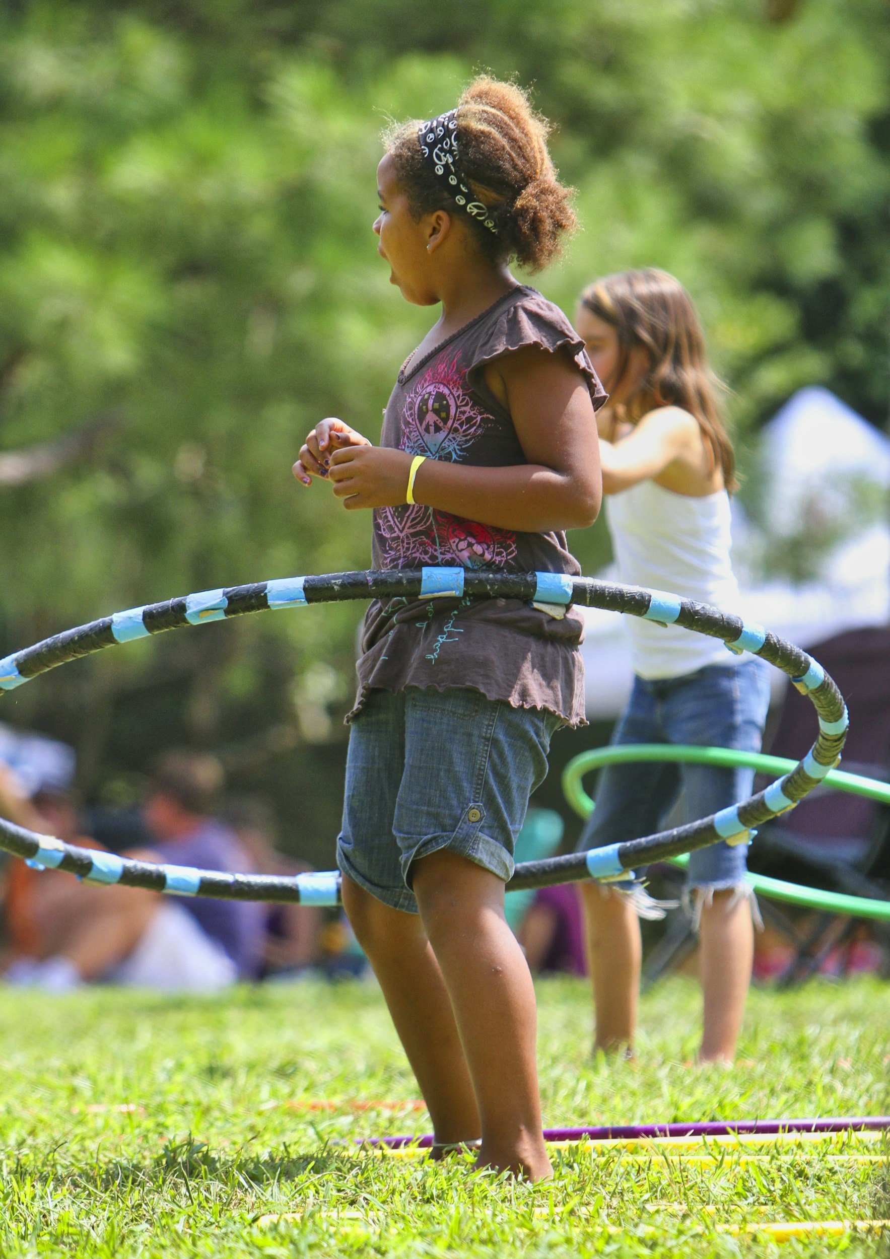 Young girl with curly hair and a black bandana style headband, standing on grass inside a hula hoop, participating in outdoor activity. She wears a t-shirt with a peace symbol and a skate shoe logo, and denim shorts. In the background, another girl with long brown hair, wearing a white top and denim shorts, is also engaged in the activity.