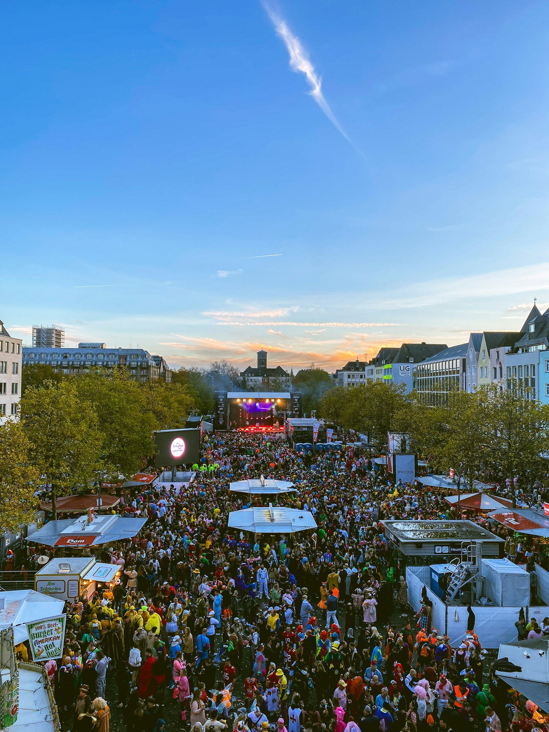 Crowd attending an outdoor concert or festival with a stage in the distance during early evening sunset with clear sky and some clouds.