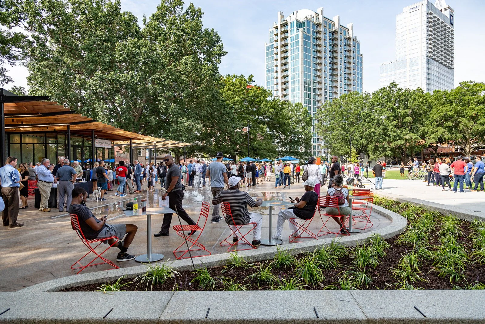 People gathered outdoors in a city park on a sunny day, some sitting at red chairs, others standing or walking, with tall buildings and lush trees in the background.