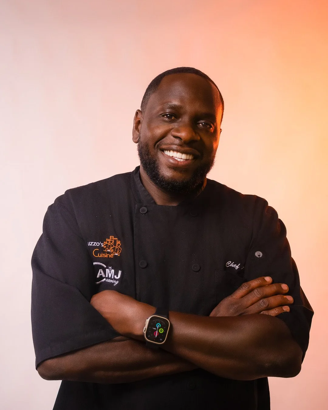 A smiling chef wearing a black chef's jacket with orange embroidery, crossed arms, and a smartwatch on his left wrist, against a light pink background.