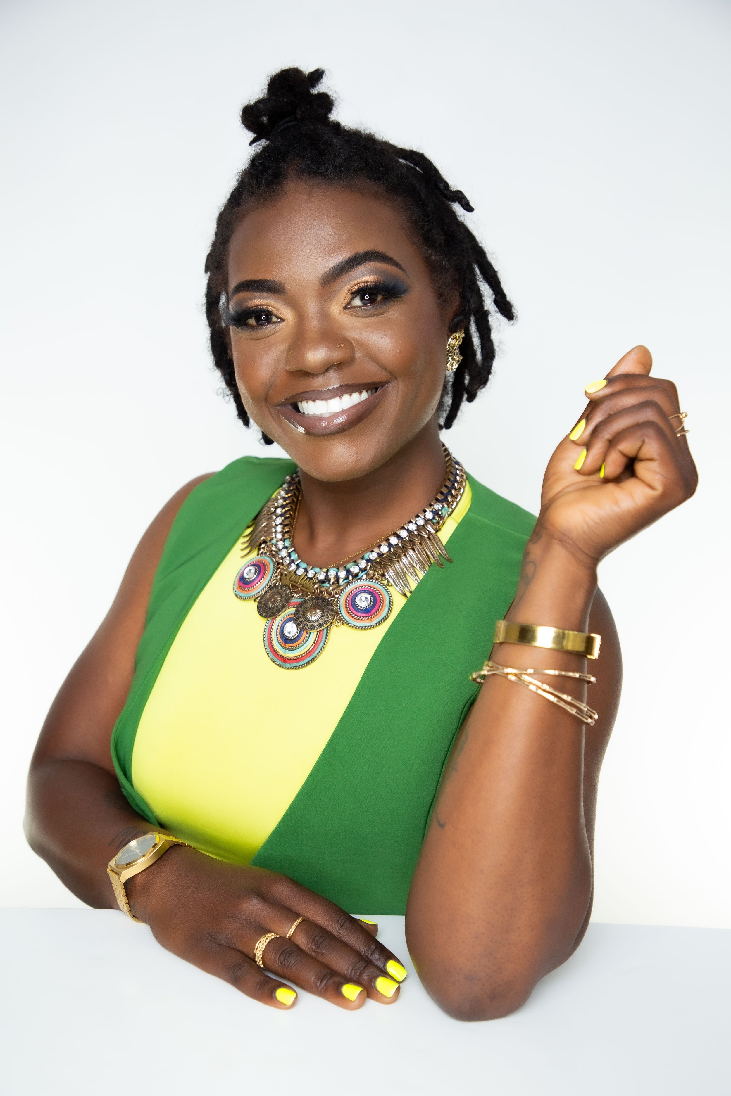 A woman with dark skin, black dreadlocks styled in a bun, and vibrant yellow nail polish, smiling confidently. She wears bold makeup, gold earrings, and layered gold jewelry, including bracelets and rings. She is dressed in a green outfit with a yellow inner lining, standing against a plain white background.