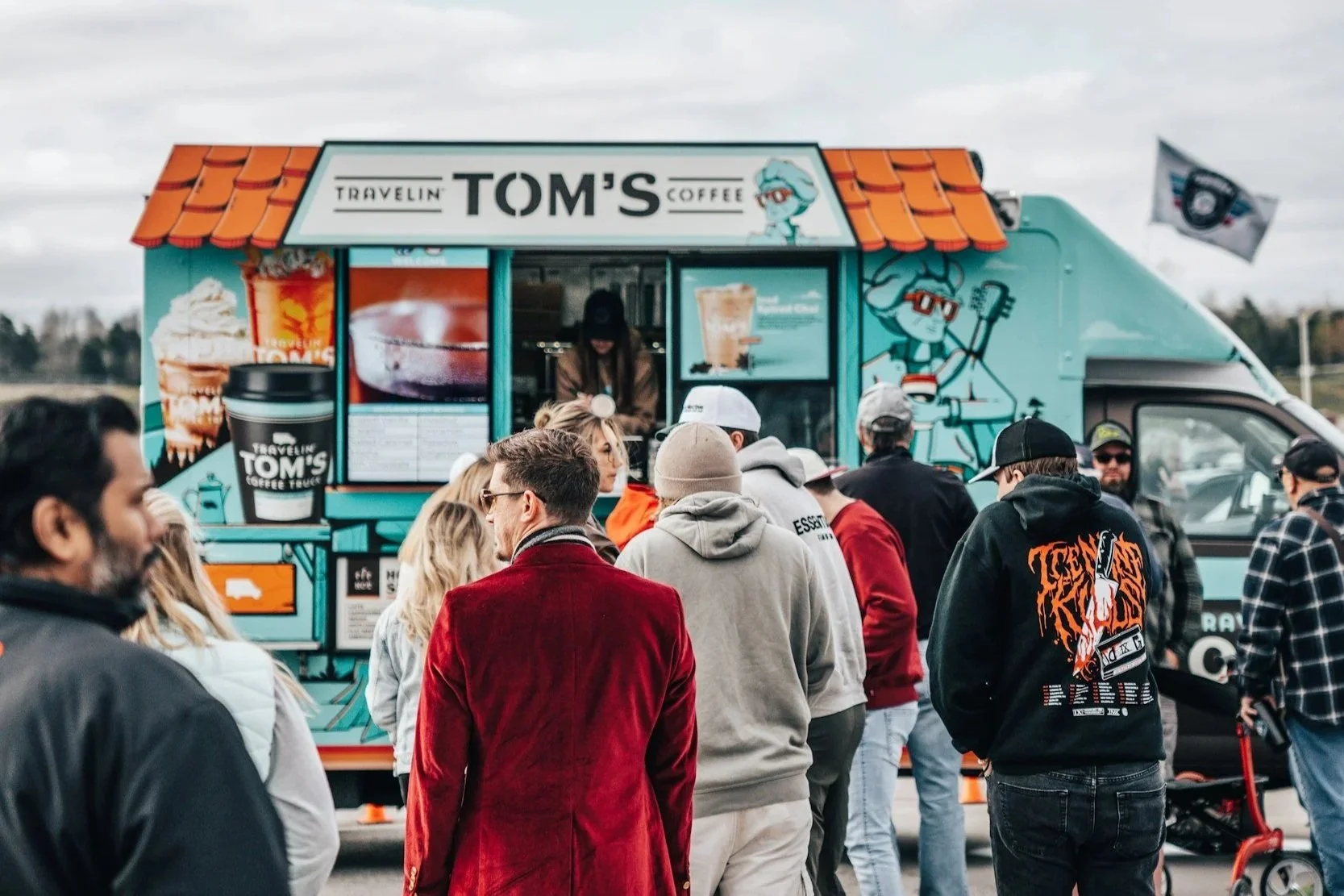 A food truck named 'Travelin Tom's Coffee' serving coffee and beverages, with a crowd of people gathered around during daytime.