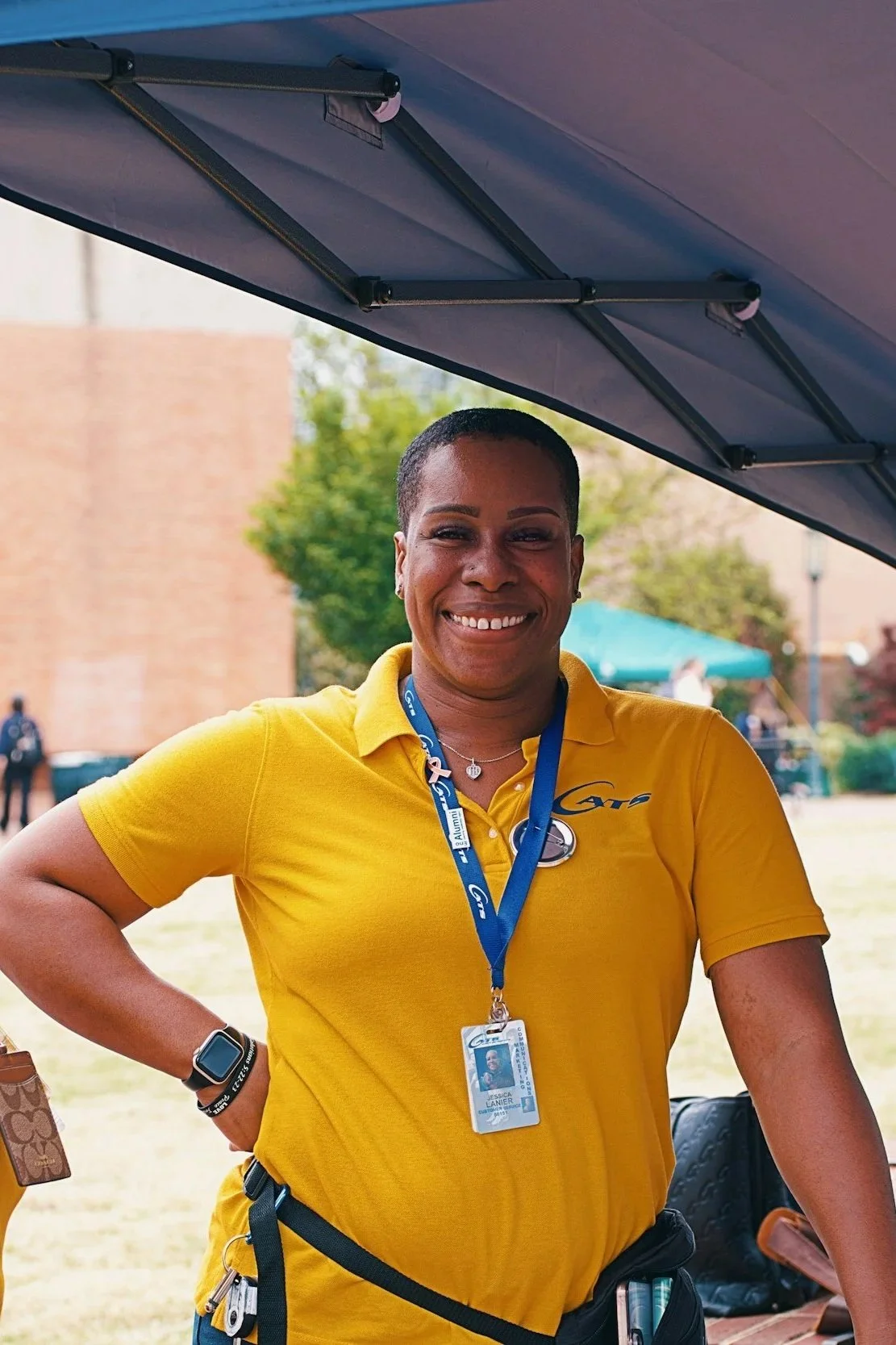 A smiling woman wearing a yellow polo shirt with a lanyard and ID badge, standing outdoors underneath a canopy.