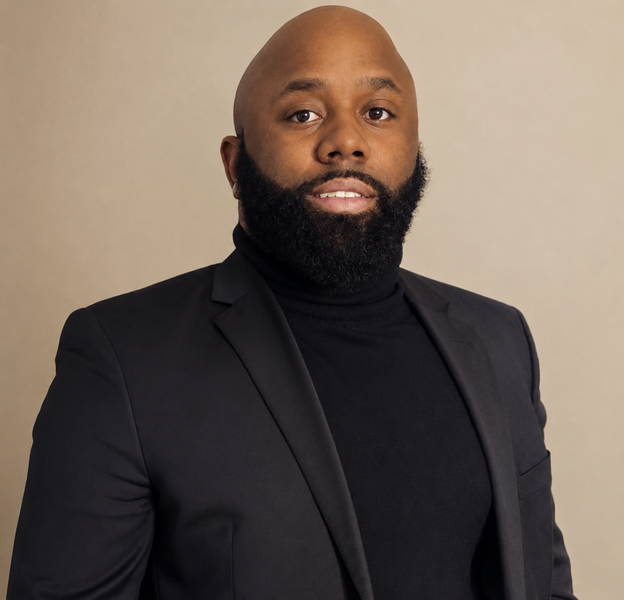 Portrait of an African American man with a bald head and full beard, wearing a black suit jacket and turtleneck, against a neutral background.
