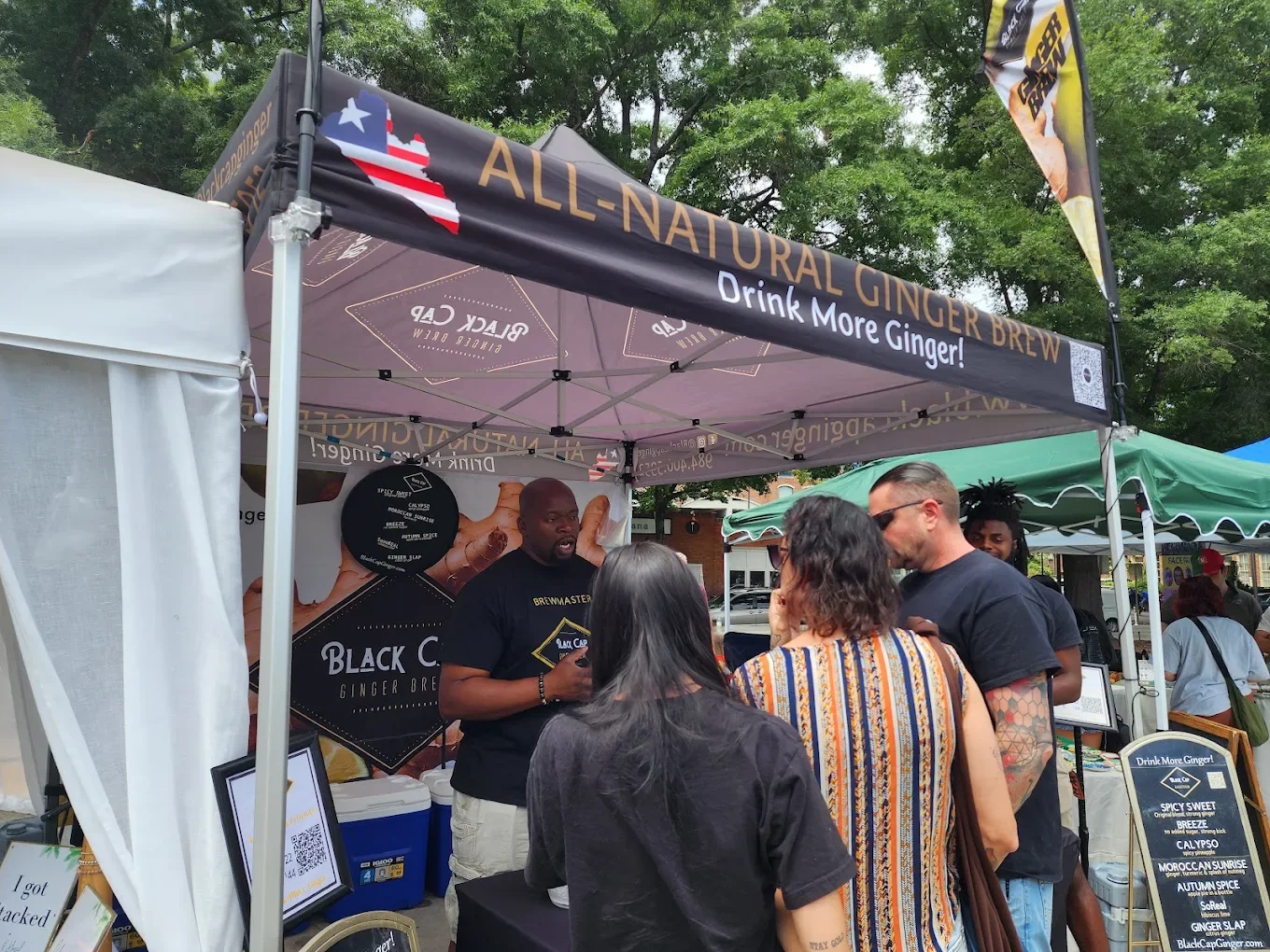 A group of people ordering ginger brew at a market stall with a black canopy that reads "All-Natural Ginger Brew" and "Drink More Ginger!" in golden letters. The stall has a banner promoting Black Cap Ginger Brew with a spicy sweet flavor. There are trees and other market stalls in the background.