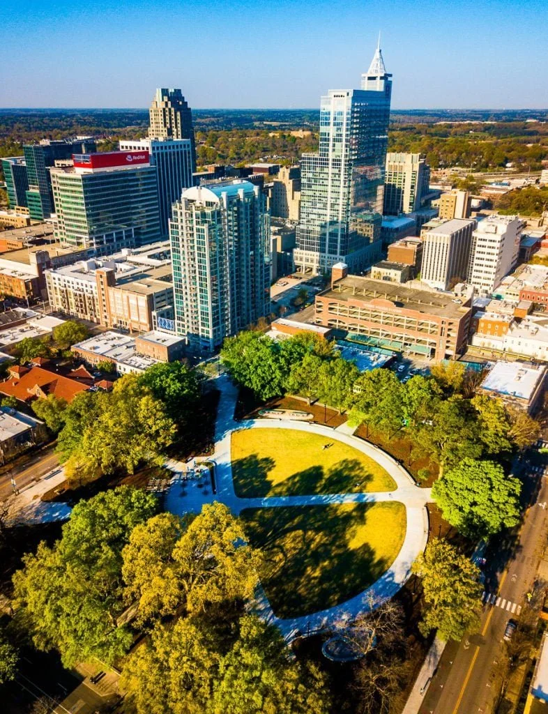 Aerial view of a cityscape with modern skyscrapers and a park with trees and a circular walking path in the foreground.