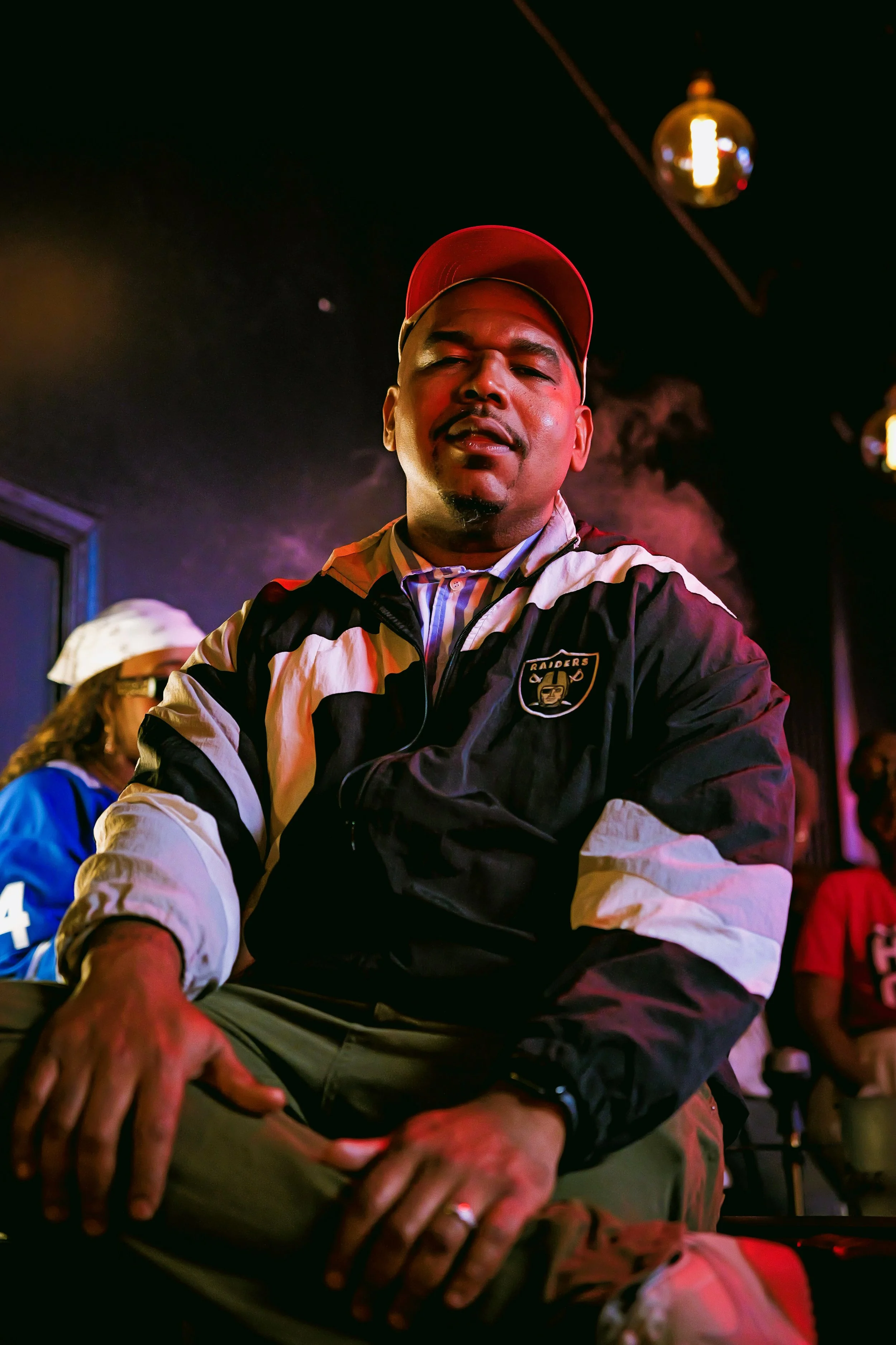 A man wearing a red cap and a black Raiders jacket sitting indoors with colored lighting and other people around.