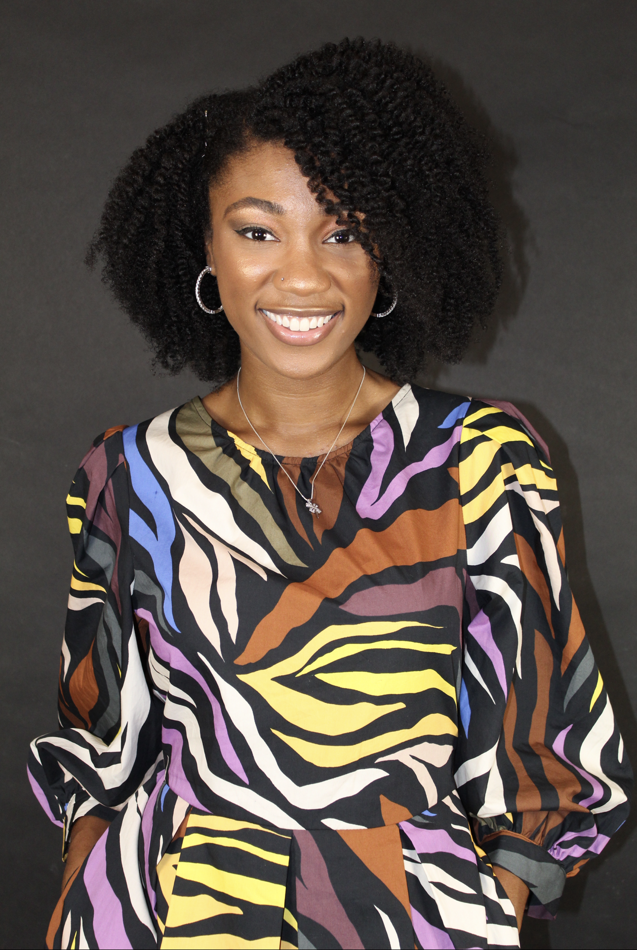 A young woman with curly black hair, wearing lipstick, jewelry, and a colorful animal print dress, smiling against a dark background.