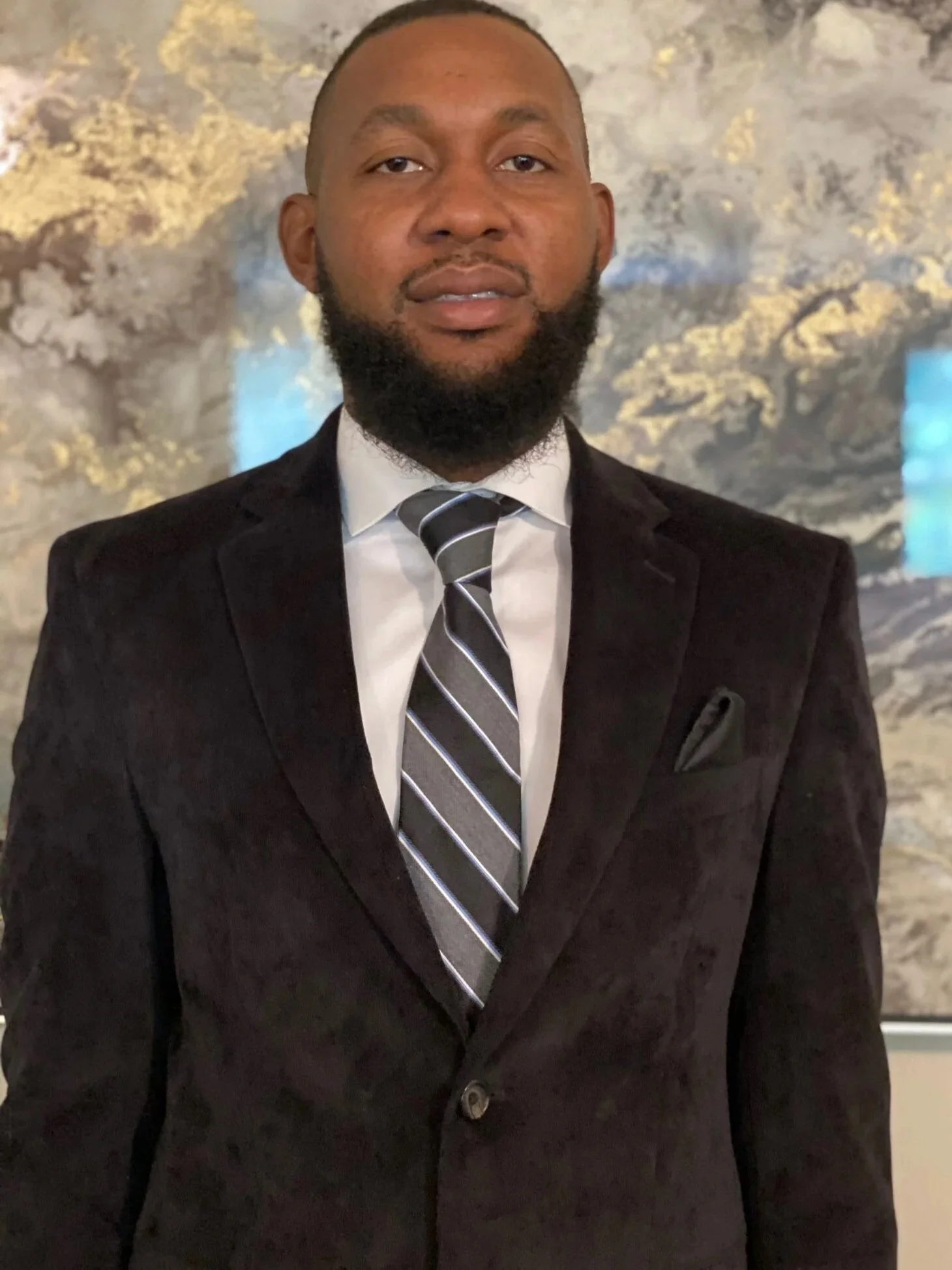 A man dressed in a black suit, white shirt, and striped tie standing in front of a textured abstract background.