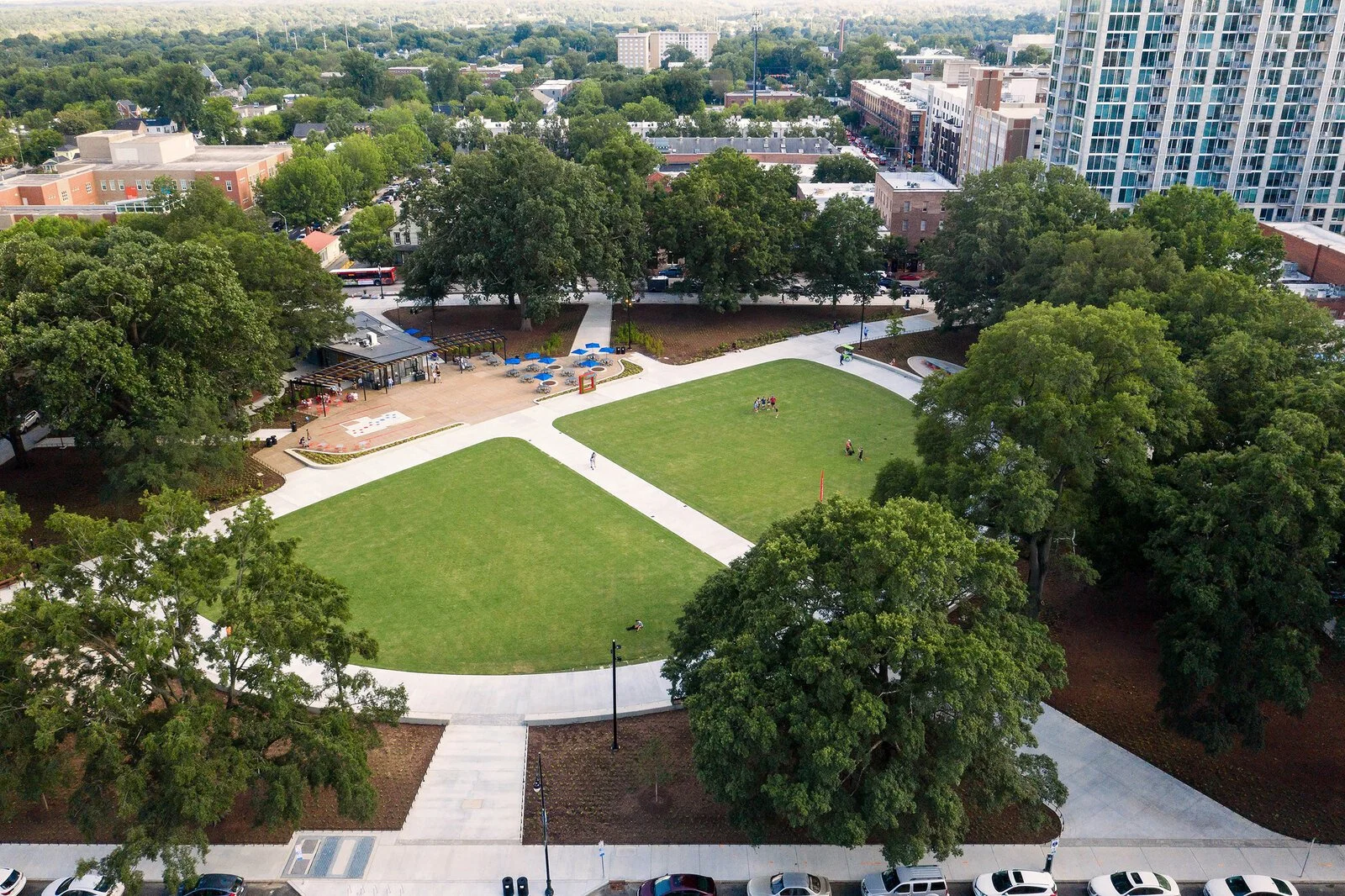 An aerial view of a park in an urban area with green lawns, pathways, trees, a playground, and surrounding buildings, including high-rise apartments and smaller structures.