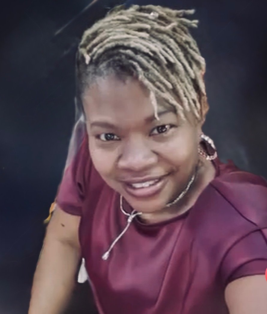 A person with short dreadlocks, smiling, wearing a burgundy shirt, earrings, and a necklace, looking up at the camera.