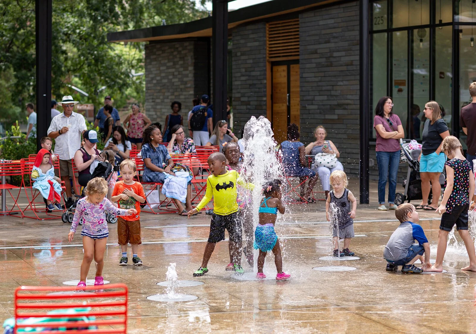 Children playing in water fountain on a town square; adults seated and standing nearby, some taking photos, some chatting.