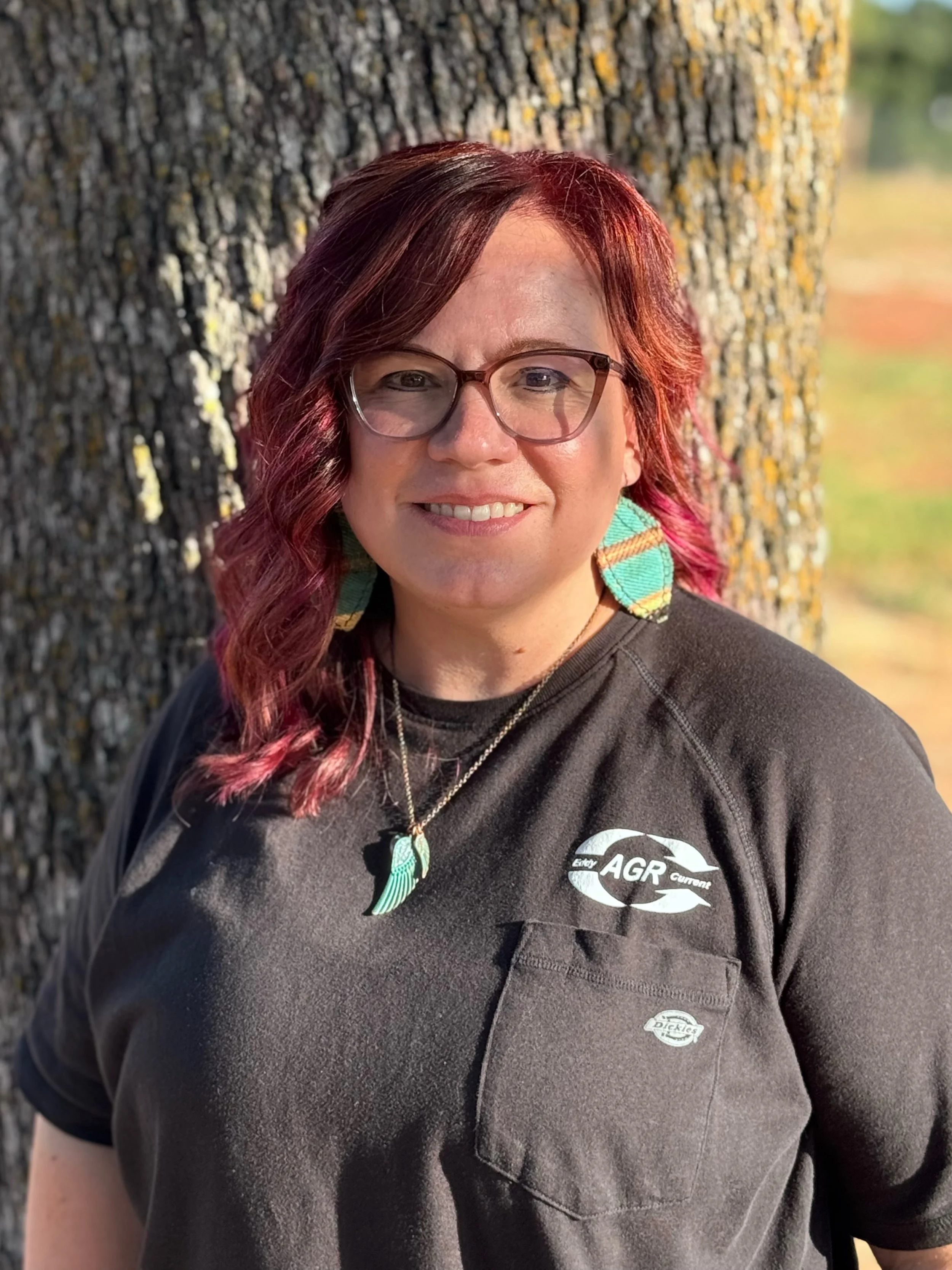 Close-up of a woman with red hair, glasses, and colorful earrings leaning against a tree outdoors.