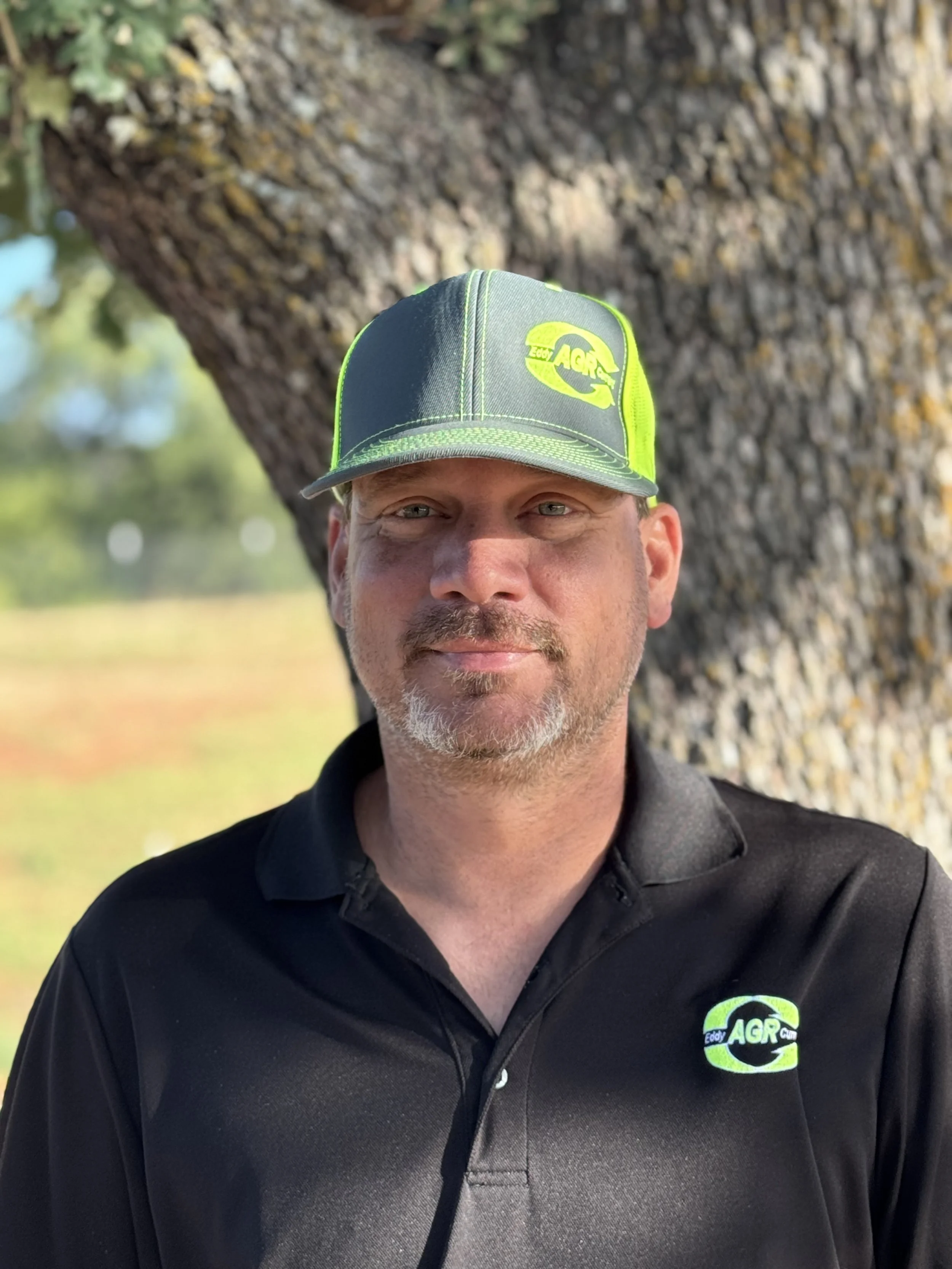 A man wearing a black polo shirt and a green and gray baseball cap stands in front of a large tree trunk, with a badge that reads 'AGR'.