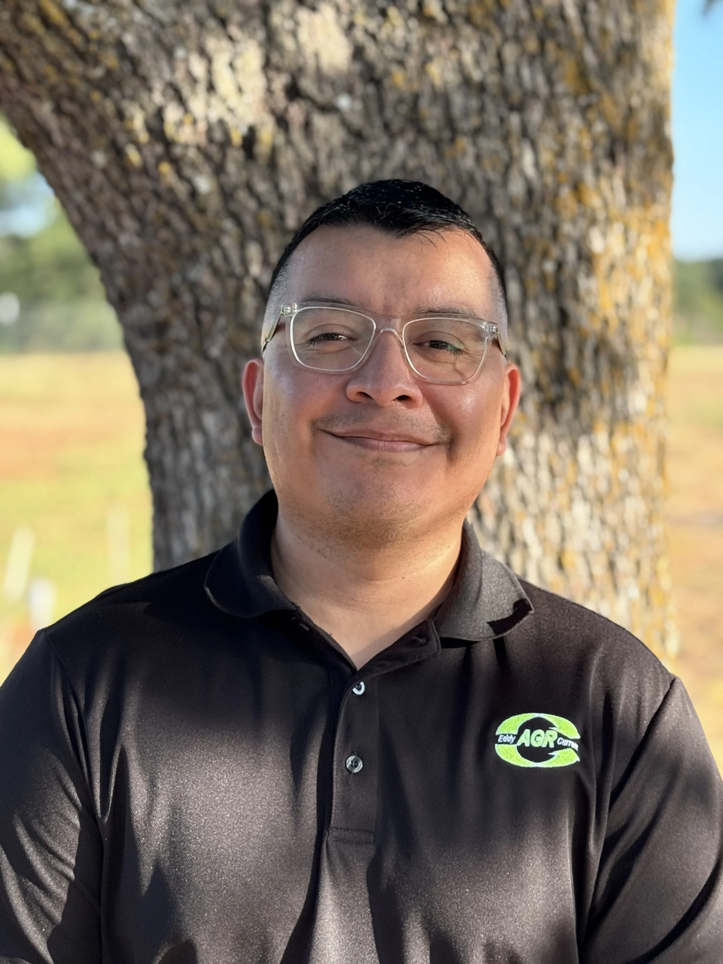 A man smiling outdoors in front of a tree, wearing glasses and a black polo shirt with an 'AGF' logo.